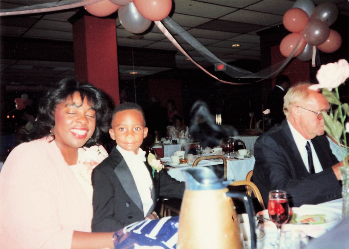 Smiling woman in pink with a young boy in a tuxedo seated at a banquet table during a decorated party with pink and gray balloons overhead.