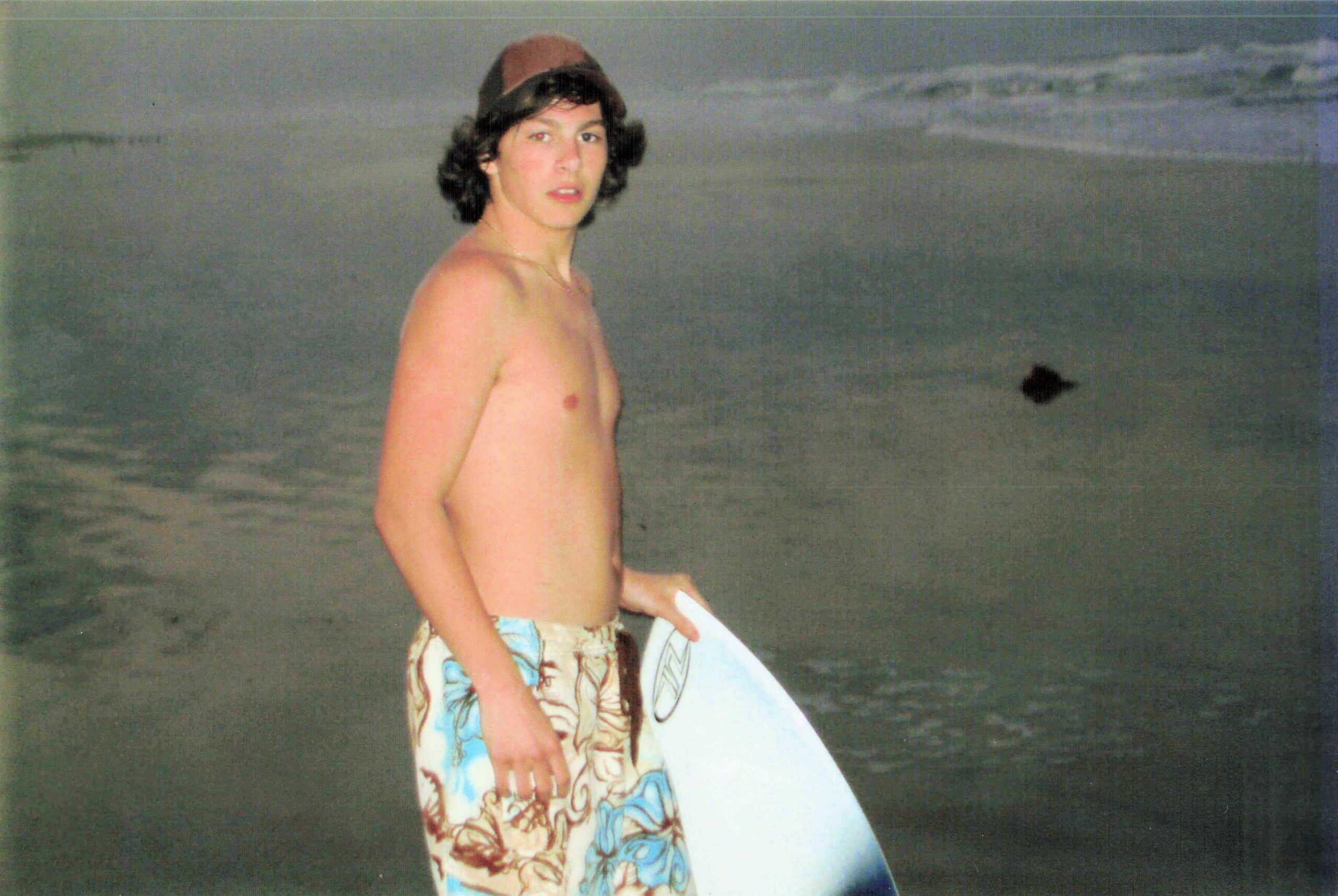 Young man shirtless on a beach, holding a light-colored surfboard with a tropical-patterned cloth draped over it, waves in the background.