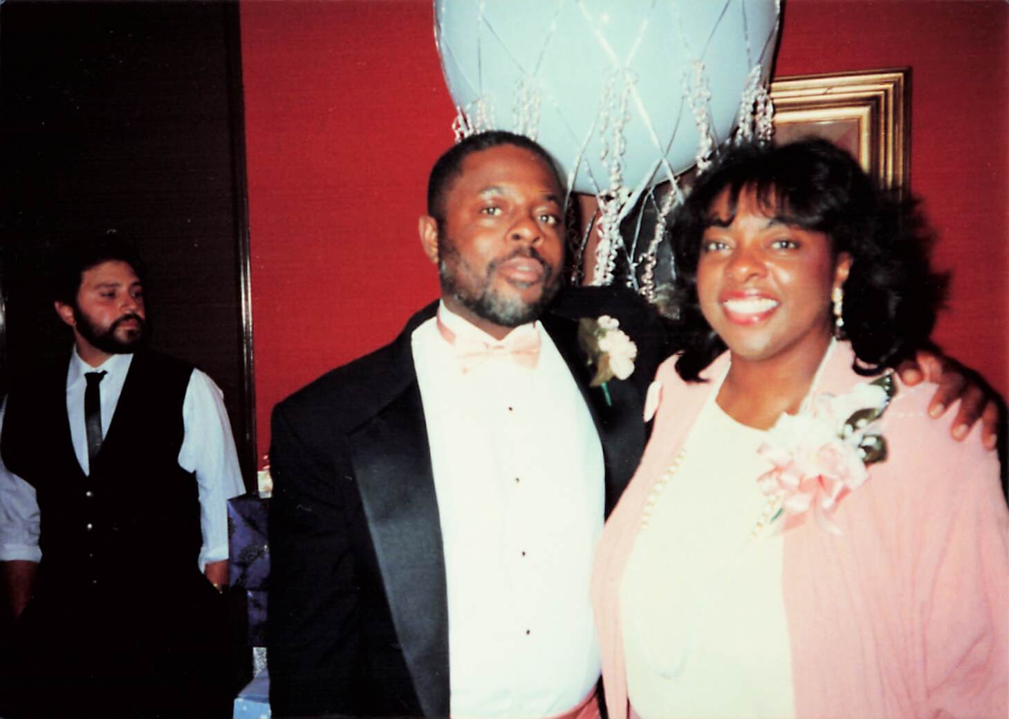 Smiling couple posing at a formal event: man in a black tuxedo with a peach bow tie and boutonniere, woman in a pink outfit with a corsage, large blue balloon in the background.