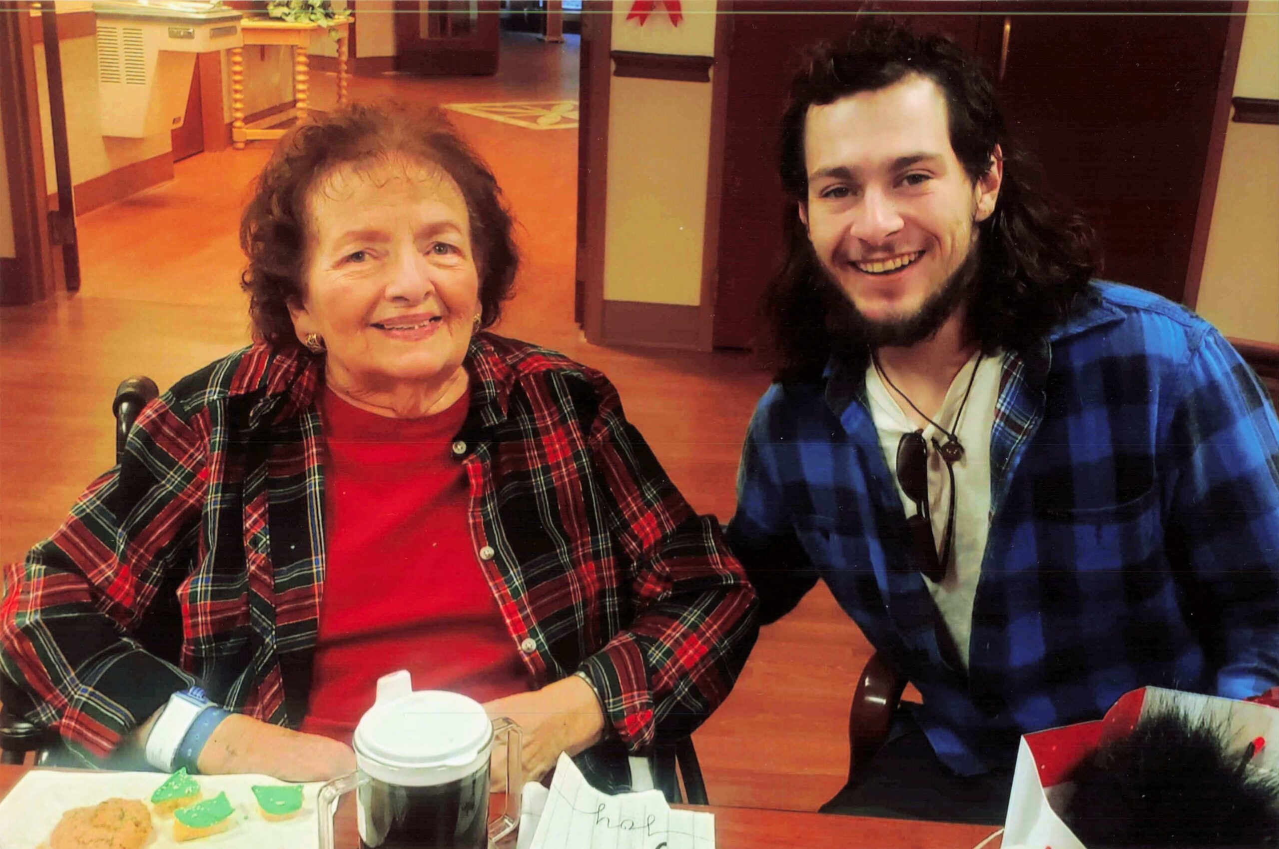 Smiling older woman in red plaid sits with a younger man in blue plaid at a cafe table, beverages and cookies visible in front of them.