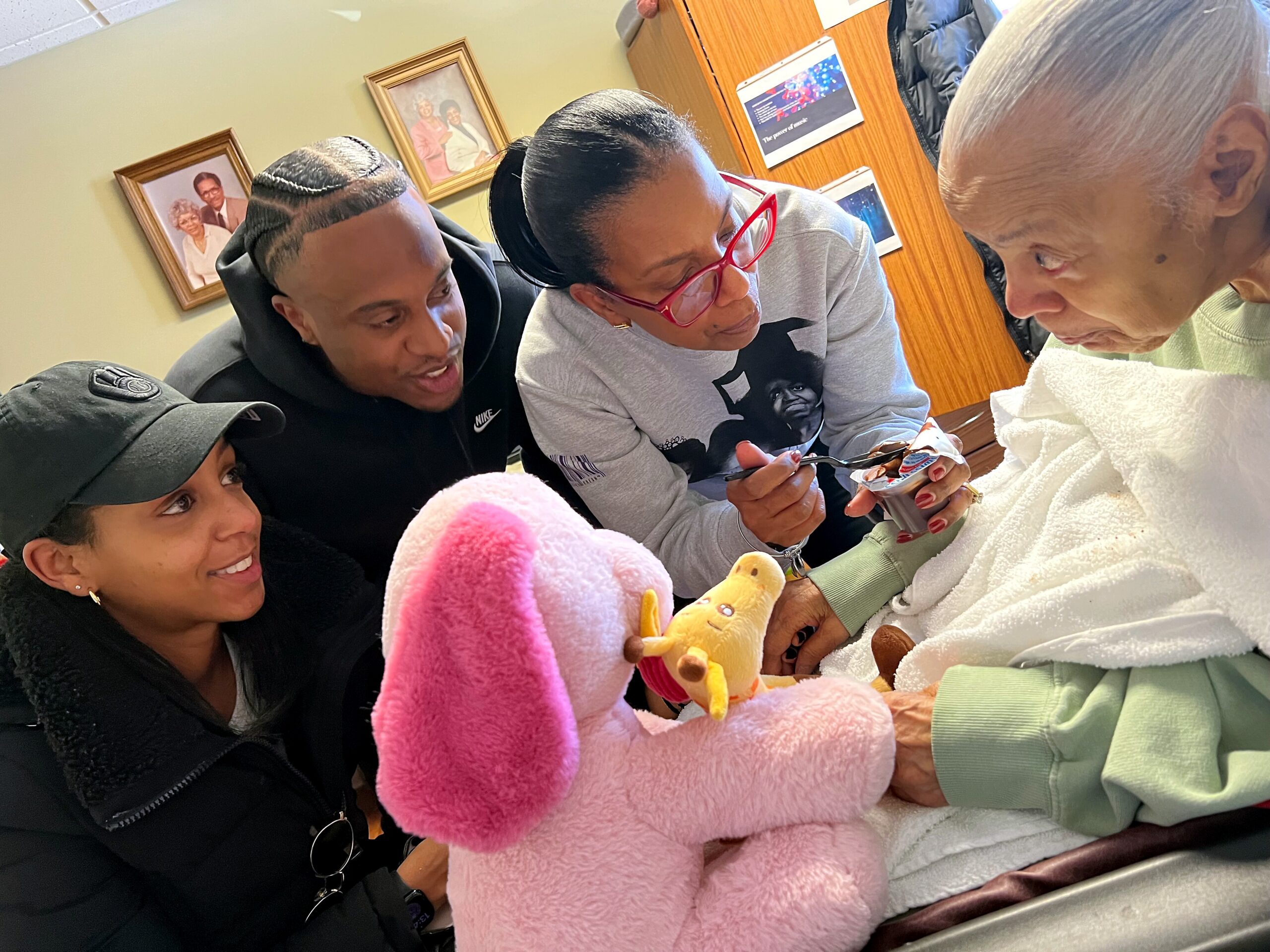 Three adults and a patient sit closely together in a hospital room, smiling at a large pink bunny stuffed toy they’re holding or showing to the patient.