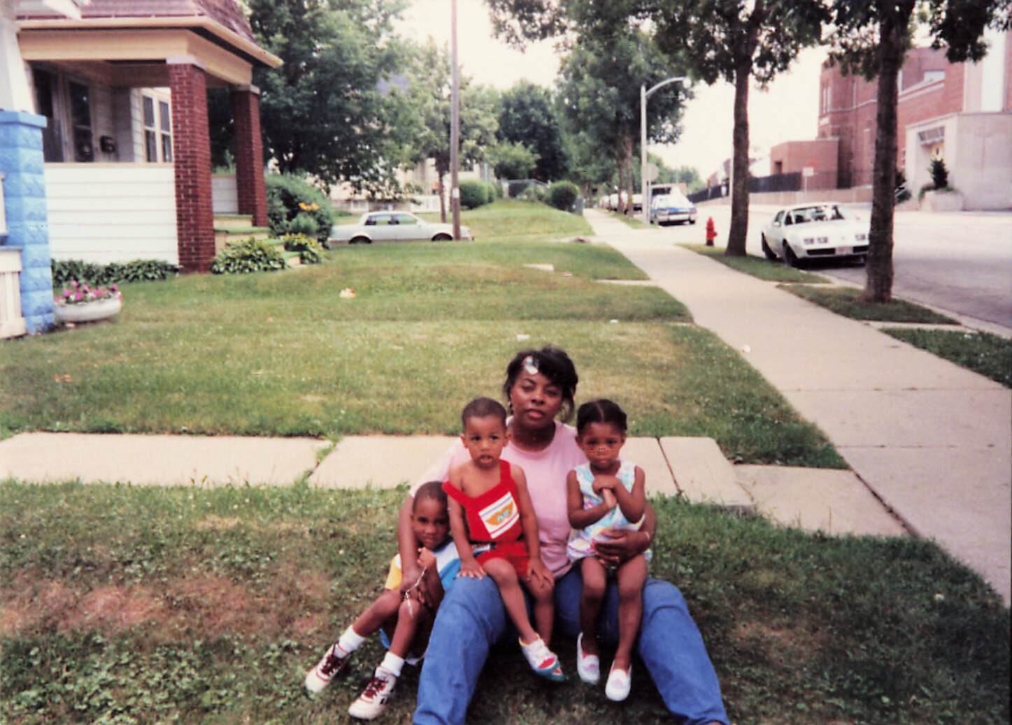 Woman sitting on the grass with three young children on a residential street; houses, trees, and parked cars in the background.