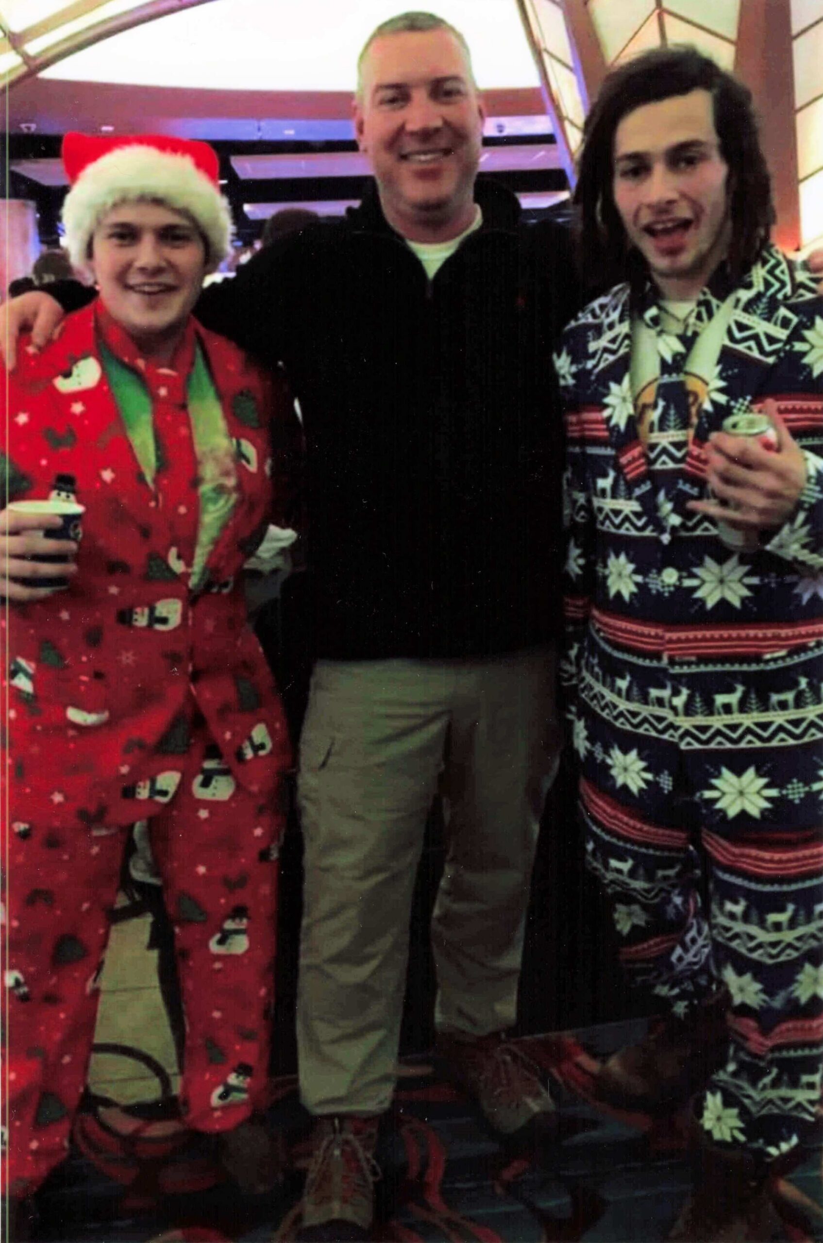 Three friends posing together at a holiday party; two wear Christmas-themed onesies (left red with Santa hat, right blue with snowflakes) and hold drinks.