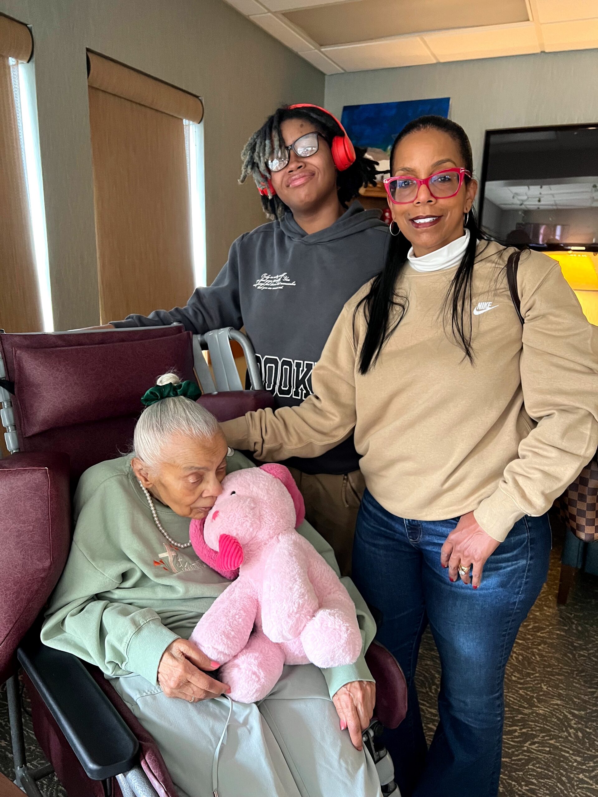 Older woman in a wheelchair holding a pink plush unicorn, with two smiling friends beside her in a cozy room.