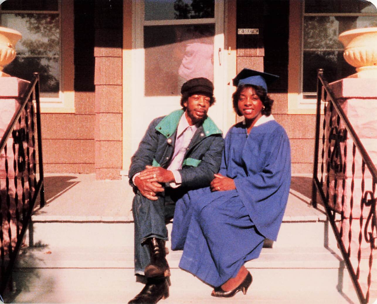 Couple sitting on the front steps of a brick house; woman in a blue graduation gown and cap, man in a jacket and cap, both smiling at the camera.