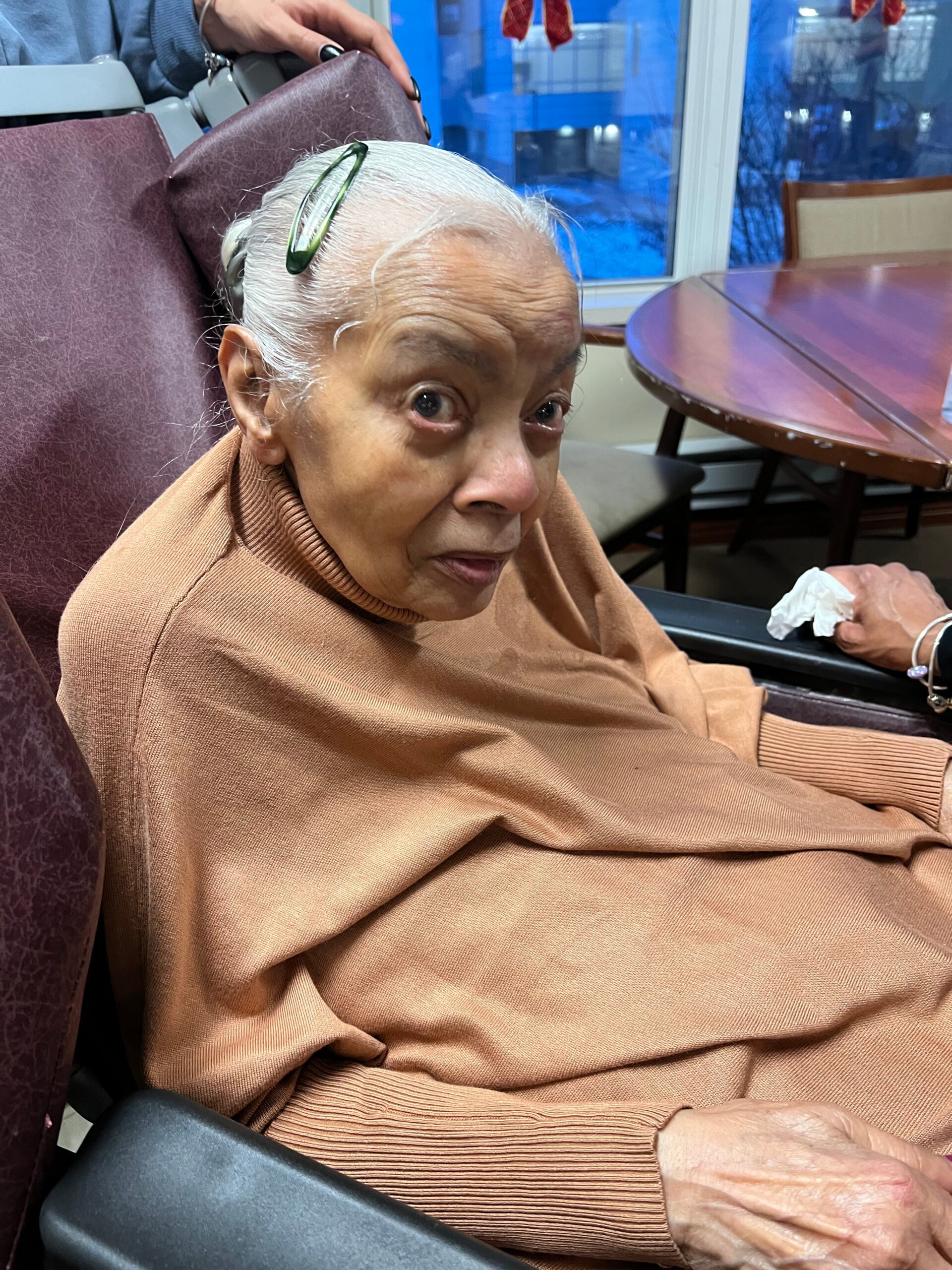 Elderly woman with white hair and green glasses resting on her head sits in a brown sweater, holding a tissue in a care setting.