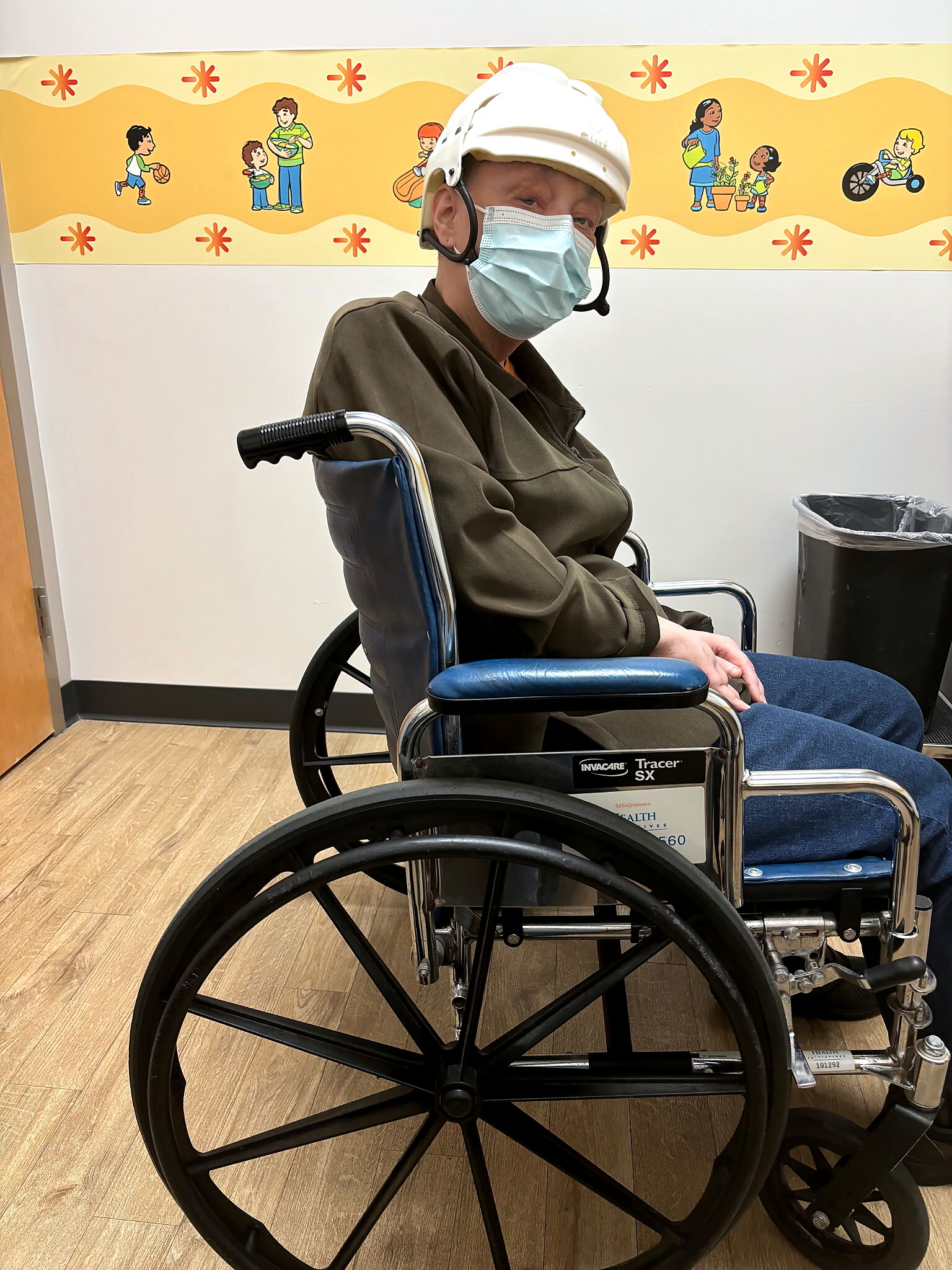 Wheelchair user wearing a white hard hat and a blue surgical mask, seated indoors with a yellow decorative wall in the background.