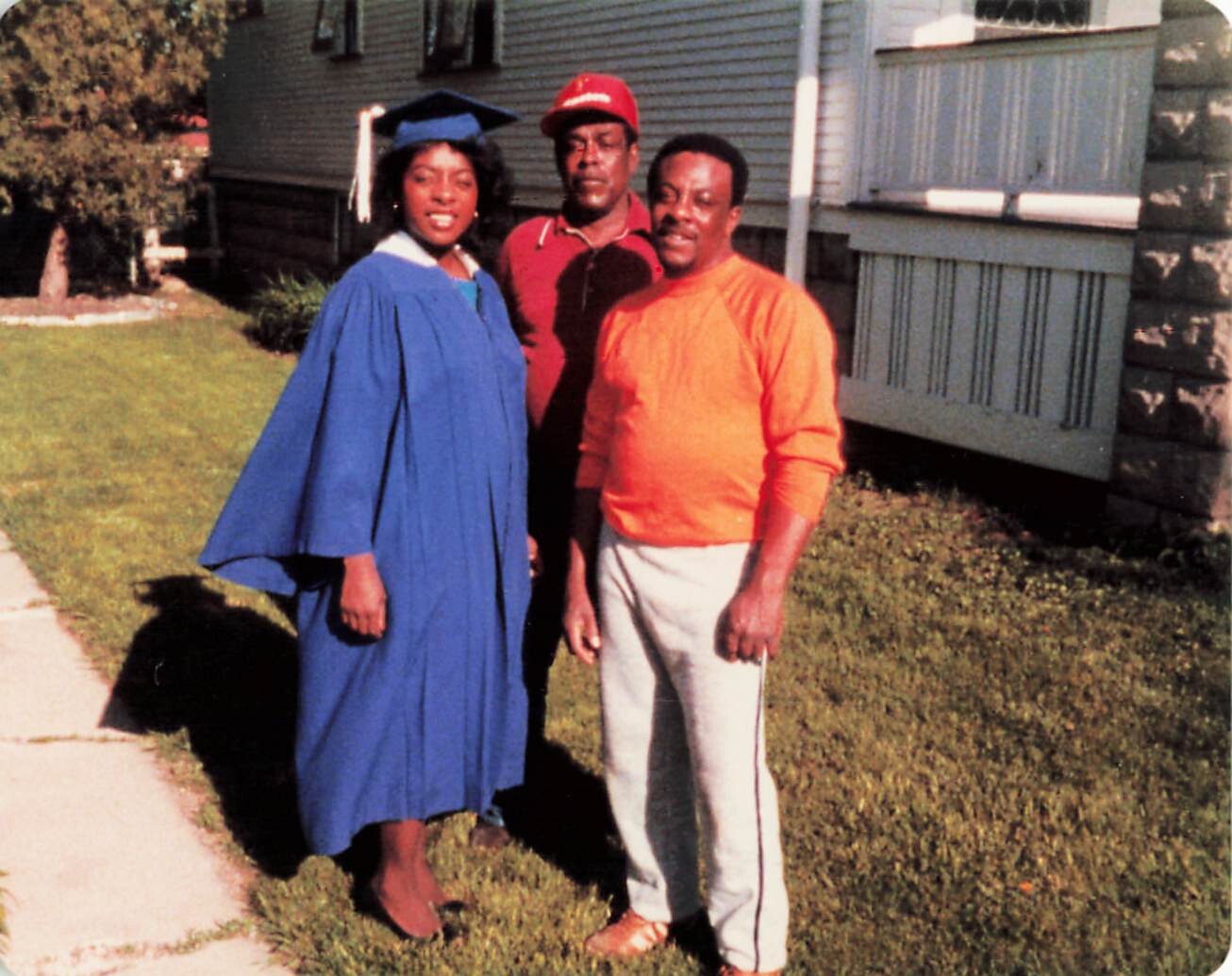 African American woman in a blue graduation gown and cap posing with two men outside a house on a sunny day.