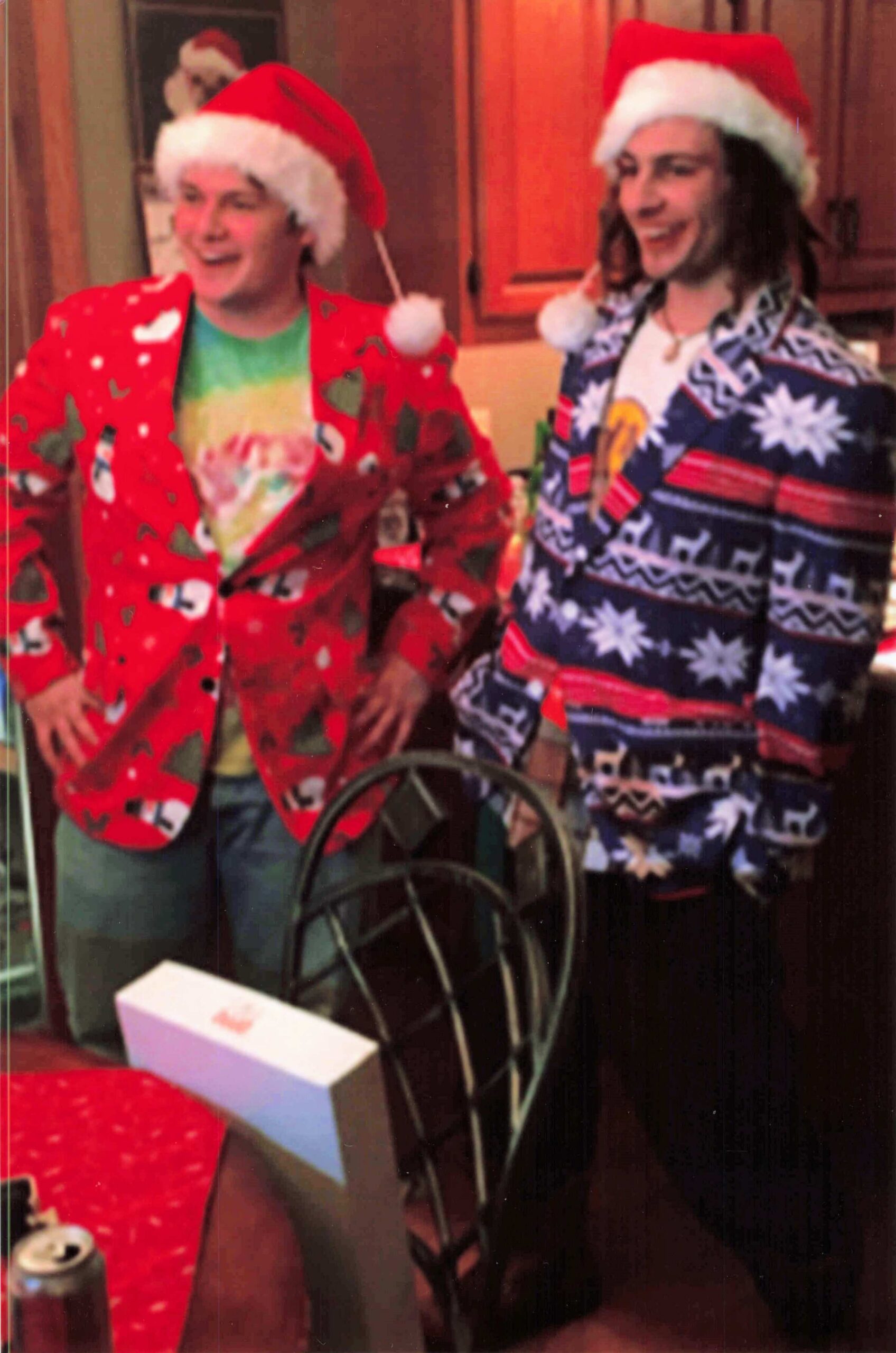 Two people wearing Santa hats and bright holiday sweaters standing indoors, smiling at a party.