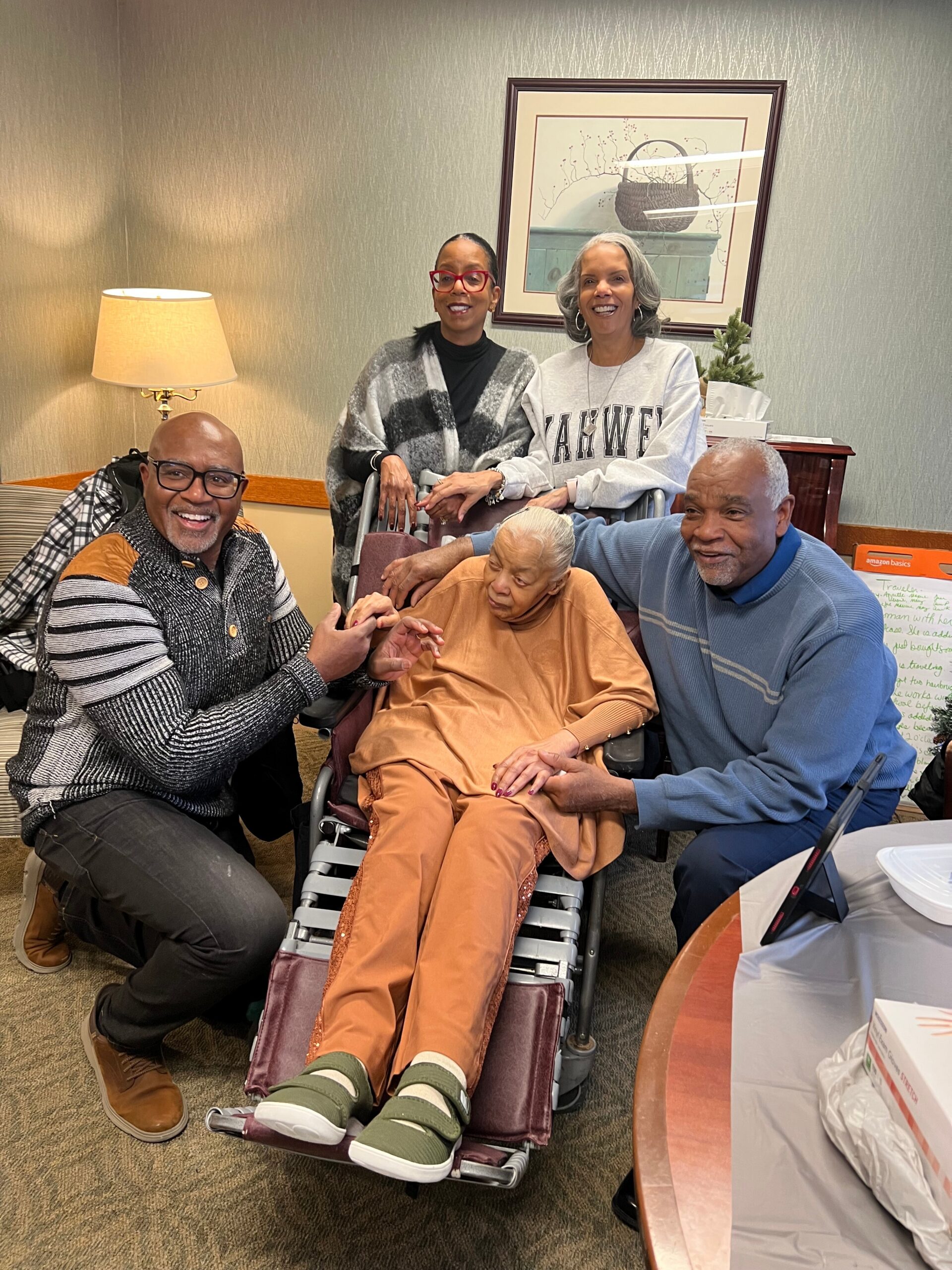 A smiling group of five adults gathered around an elderly woman in a wheelchair in a cozy room.