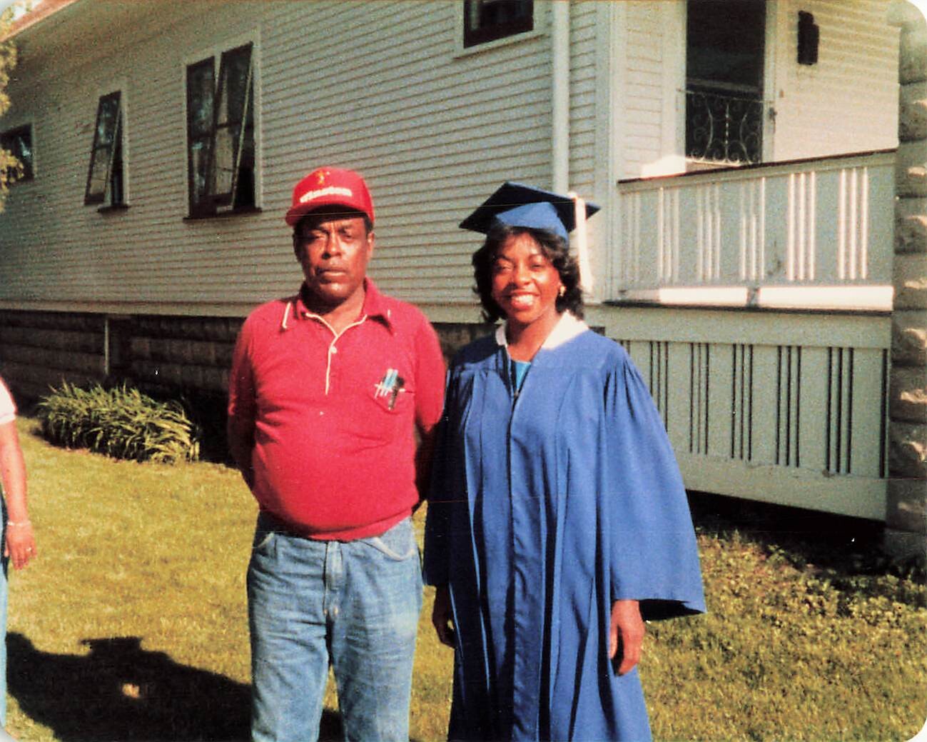 Smiling graduate in a blue cap and gown posing with a man in a red shirt and cap outside a house.