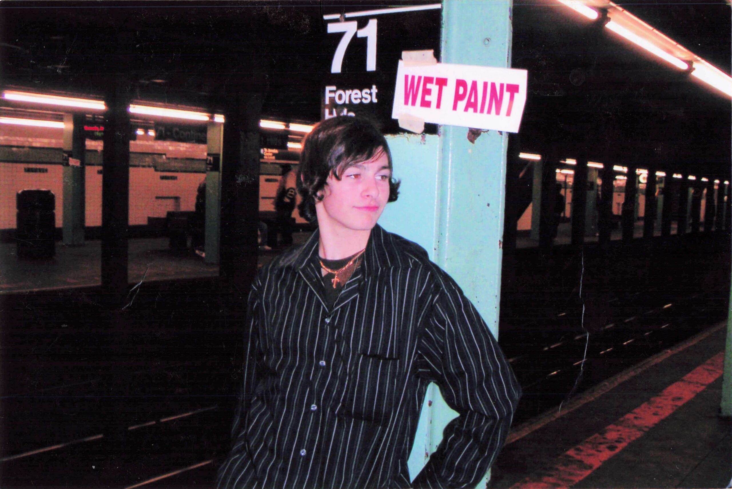 Person in a striped shirt leaning against a turquoise pillar under a sign that reads 'WET PAINT' on a subway platform at night.