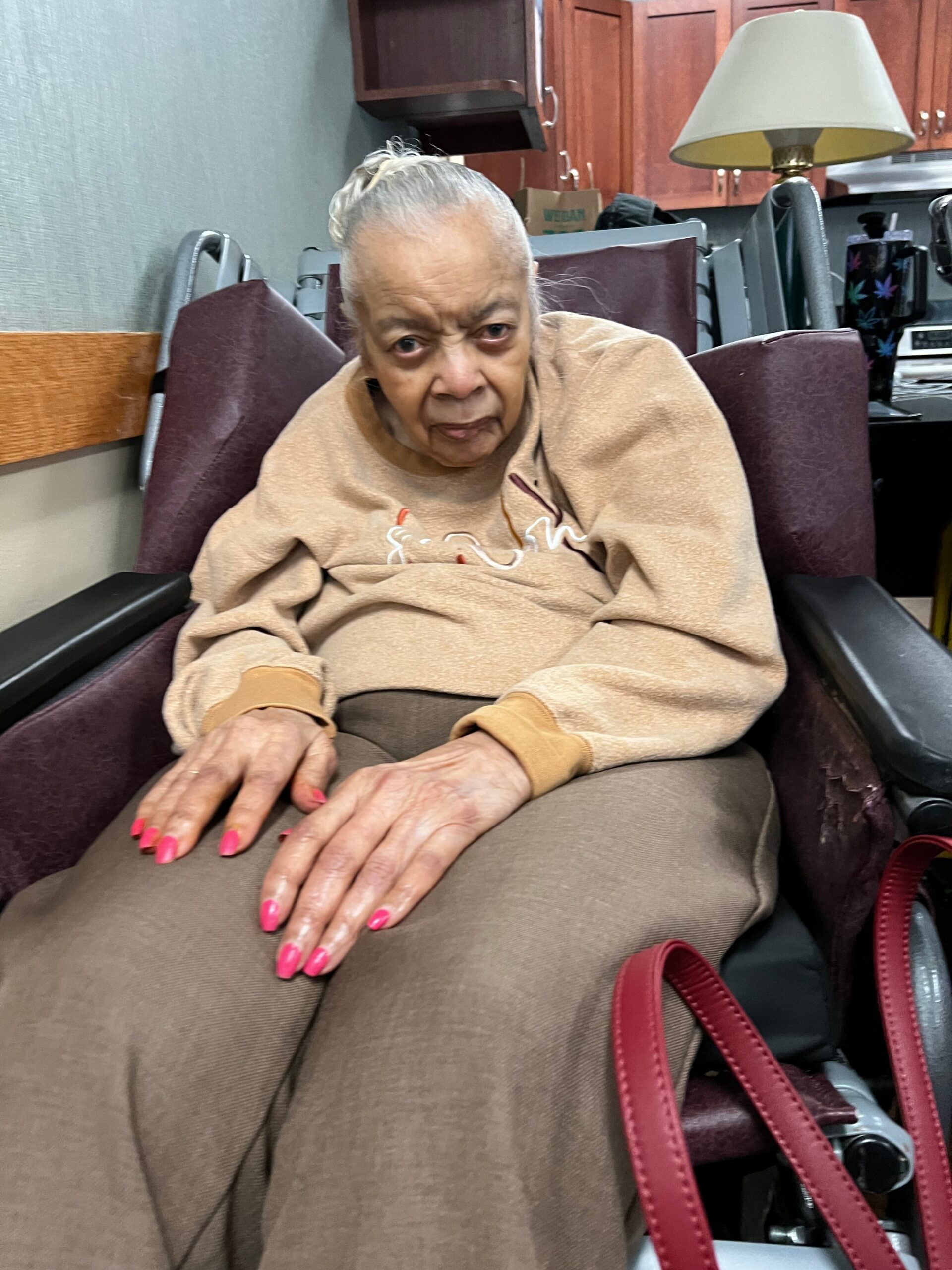 Older woman sitting in a wheelchair wearing a beige fleece, with bright pink nails visible on her hands. She looks toward the camera in a room with wooden cabinets and a lamp behind her.