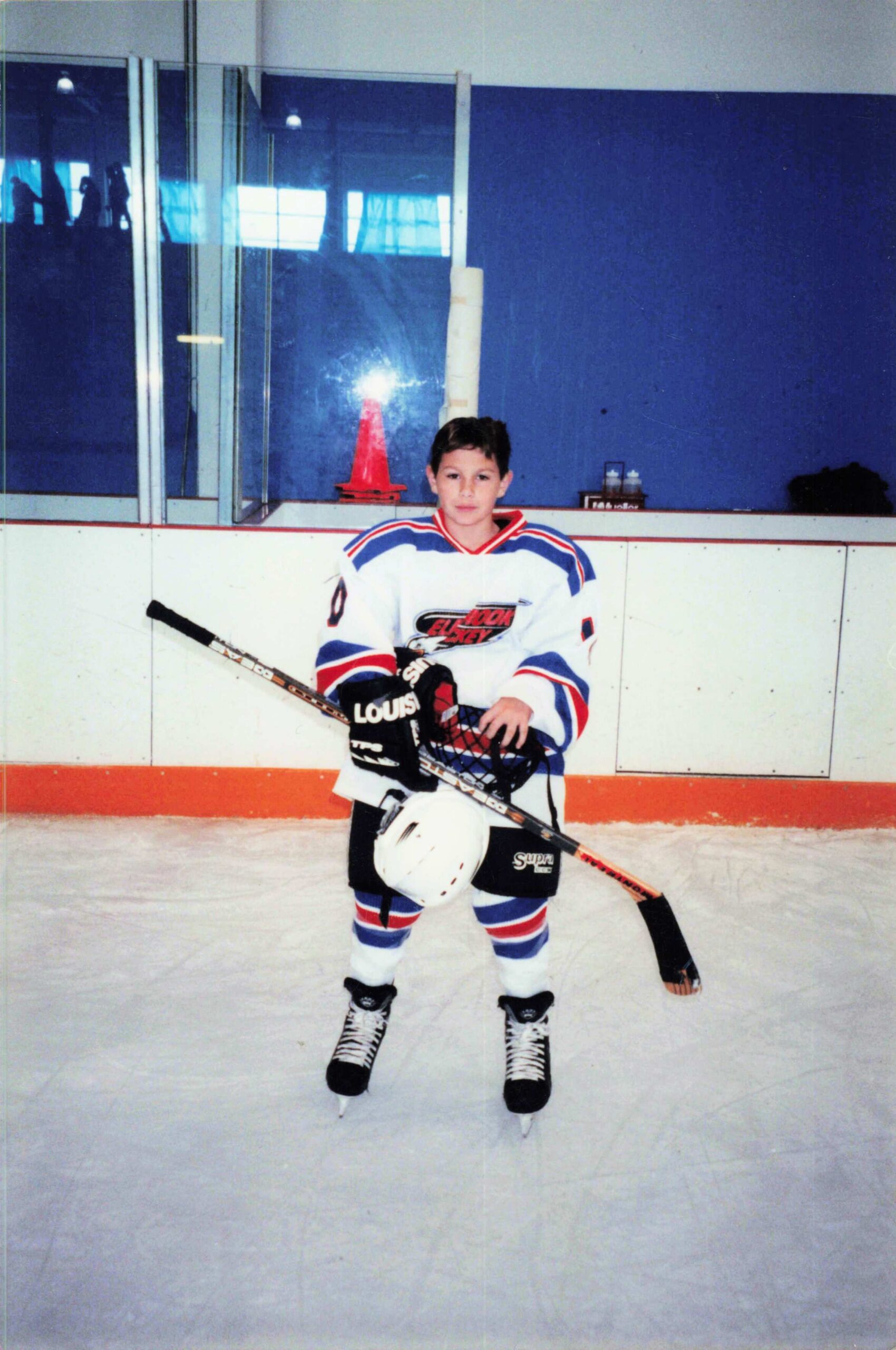 Young hockey player posing on the ice in a rink, wearing a white jersey with red and blue stripes and holding a hockey stick.