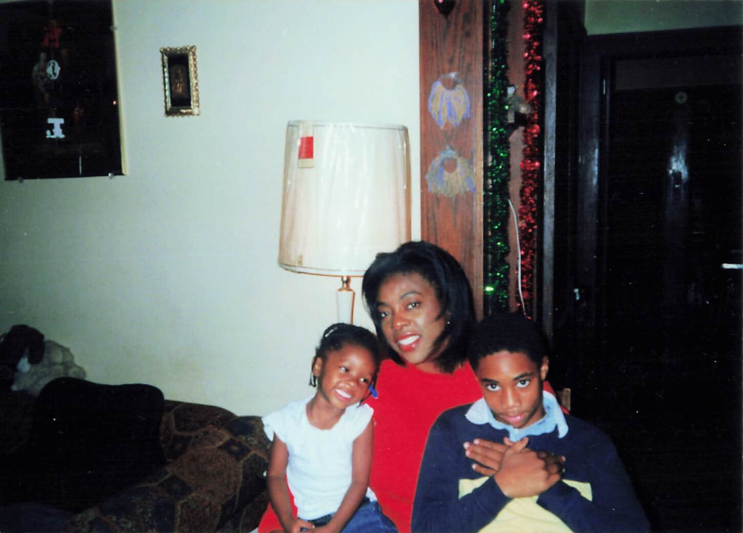 A woman in a red top sits with two children on a couch in a sparsely decorated living room during a festive season.