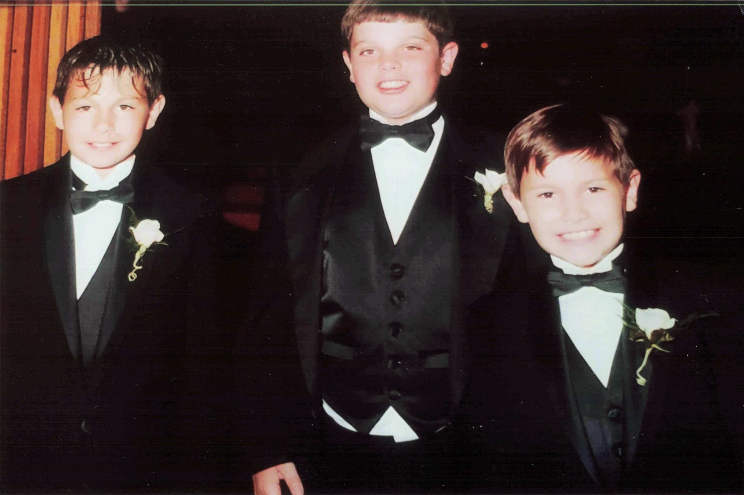 Three boys in black tuxedos with white bow ties posing for a formal photo, each wearing a boutonniere.