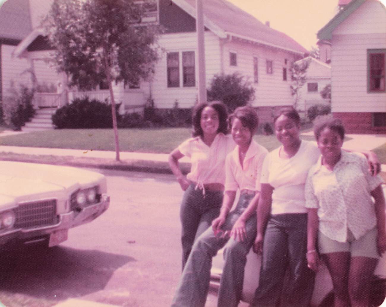 Four women pose together in front of a vintage car on a residential street, smiling at the camera.
