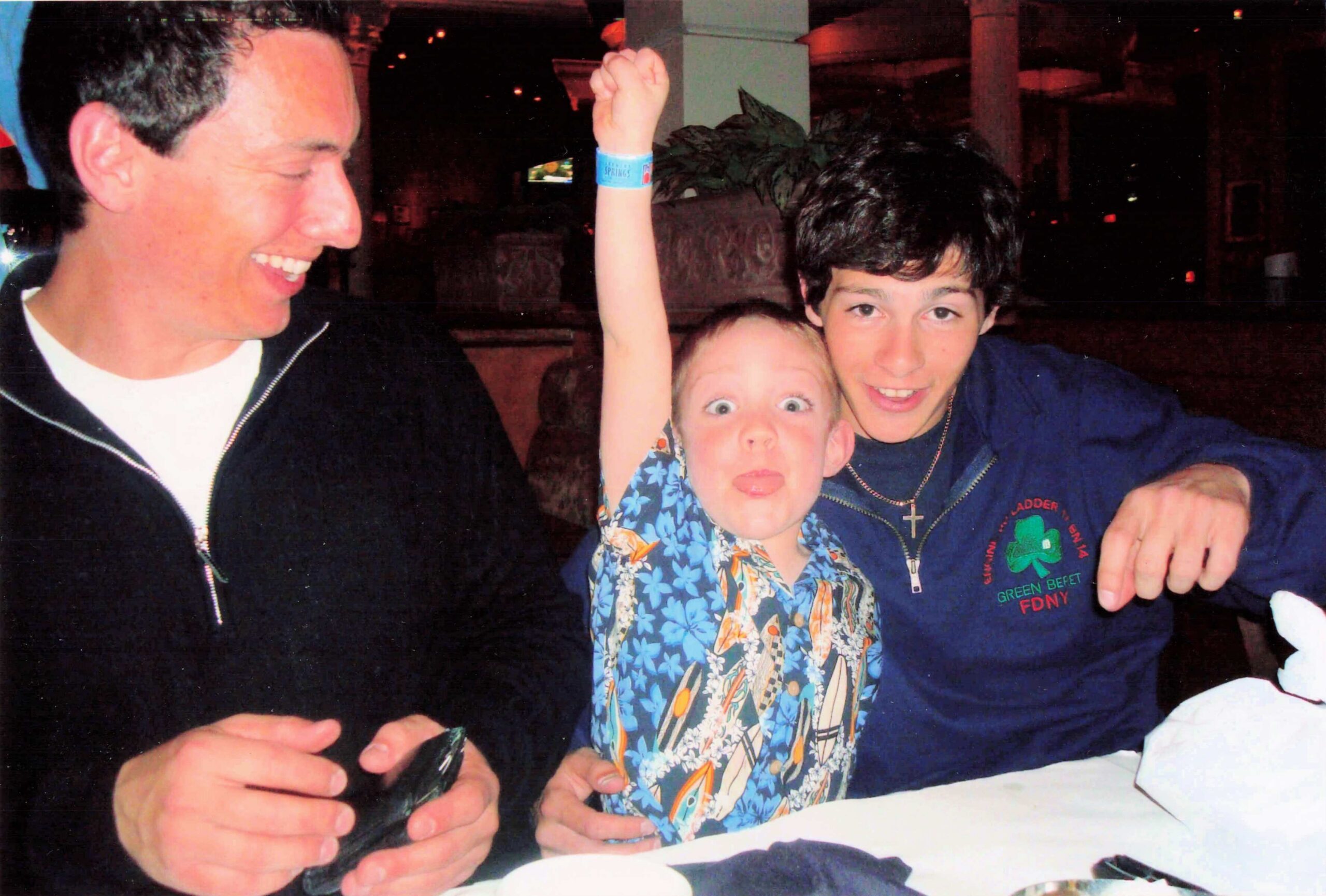 Three people at a table posing for a photo: a smiling man on the left, a child in the middle raising an arm, and a teen on the right in a blue hoodie with a necklace.