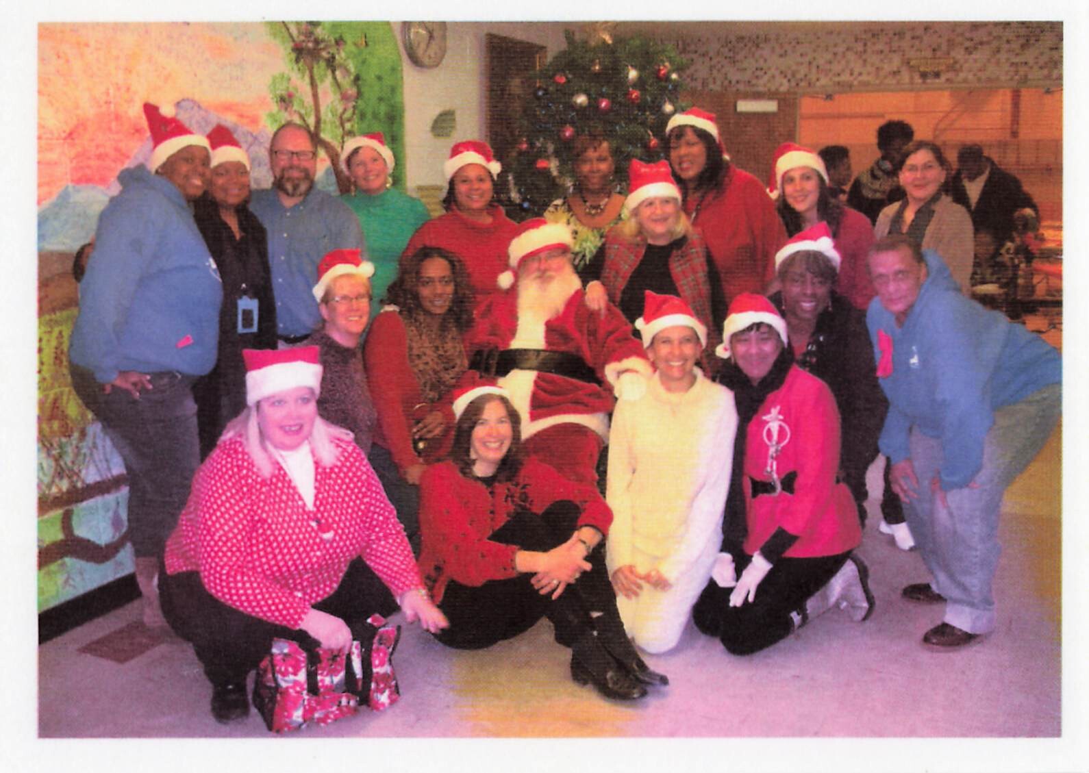 Group of adults and children in Santa hats posing with Santa beside a Christmas tree in a festive room.