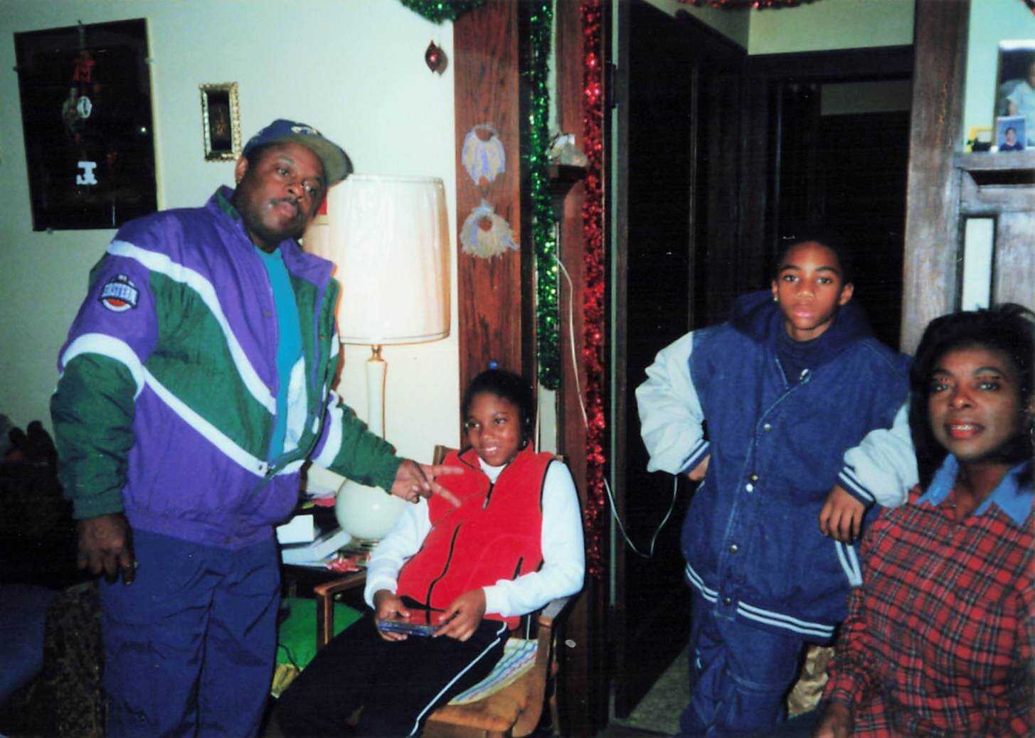 Four people posing in a festively decorated living room: a man in a colorful jacket, a girl in a red vest seated, and two teens in blue jackets standing nearby.