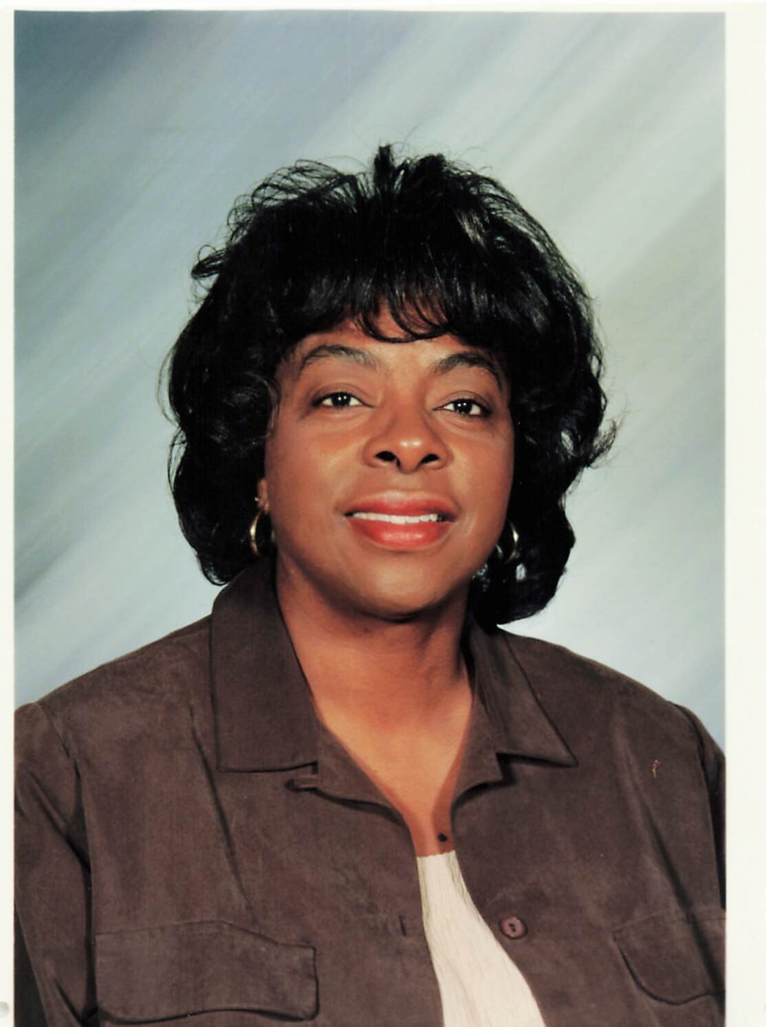 Portrait of a smiling Black woman with short black hair, wearing a brown jacket and gold hoop earrings, against a light gray backdrop.