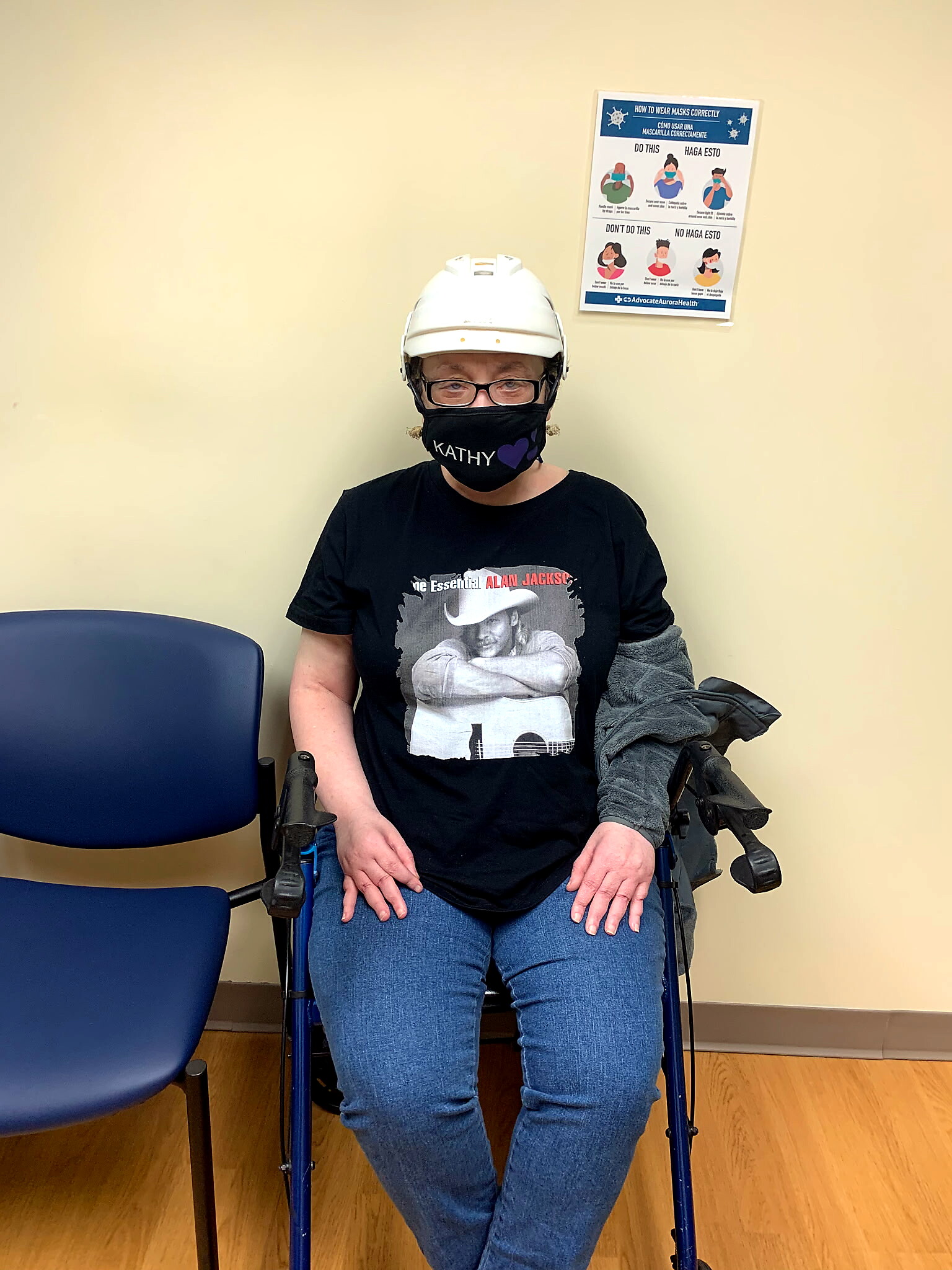 Person in a wheelchair wearing a white helmet and a black mask that reads 'KATHY' with a purple heart, indoors near a blue chair.