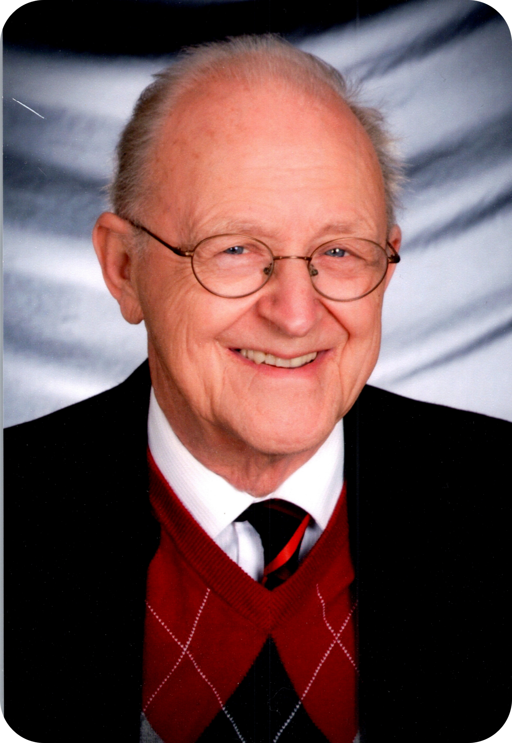 Older man with round glasses smiling, wearing a red argyle sweater and dark blazer.