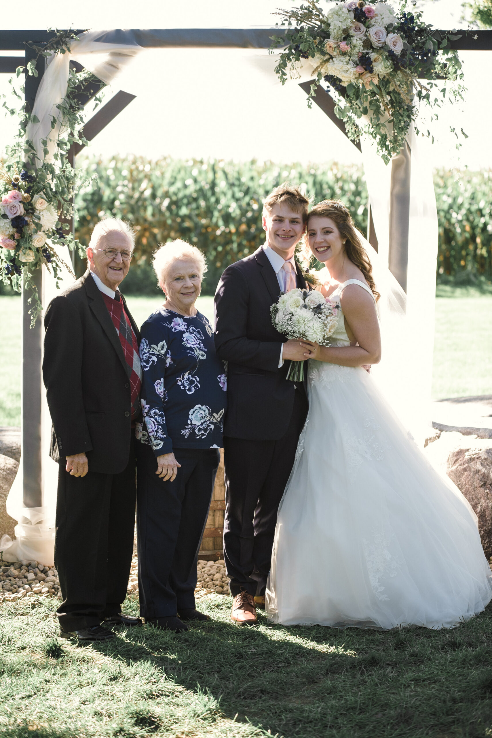 Bride and groom pose with their grandparents under a floral wedding arch outdoors, all smiling.”