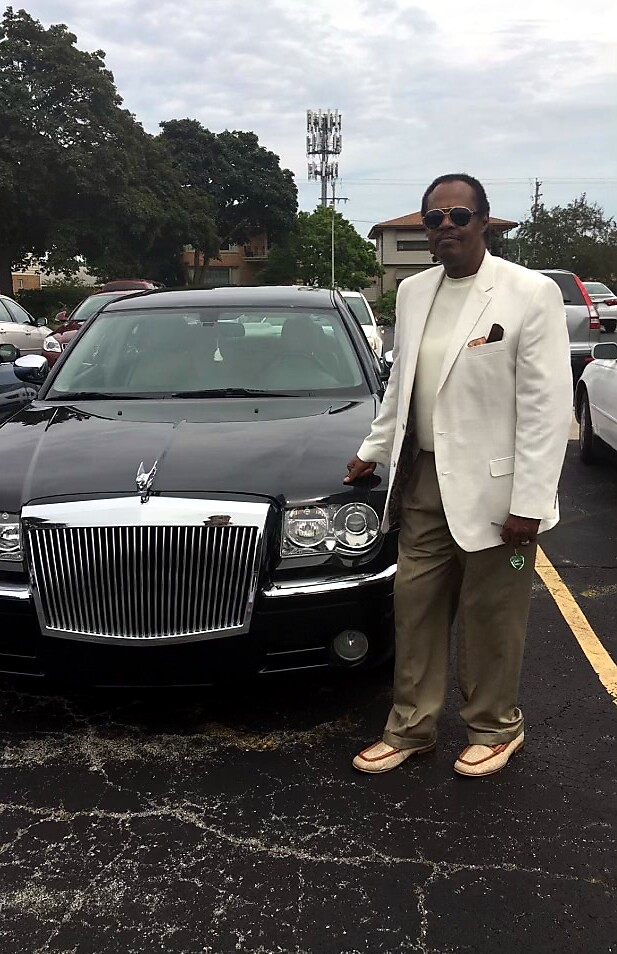 Man in a white blazer, tan trousers, and loafers stands beside a black luxury car (Rolls-Royce) in a parking lot, wearing sunglasses and a pocket square.