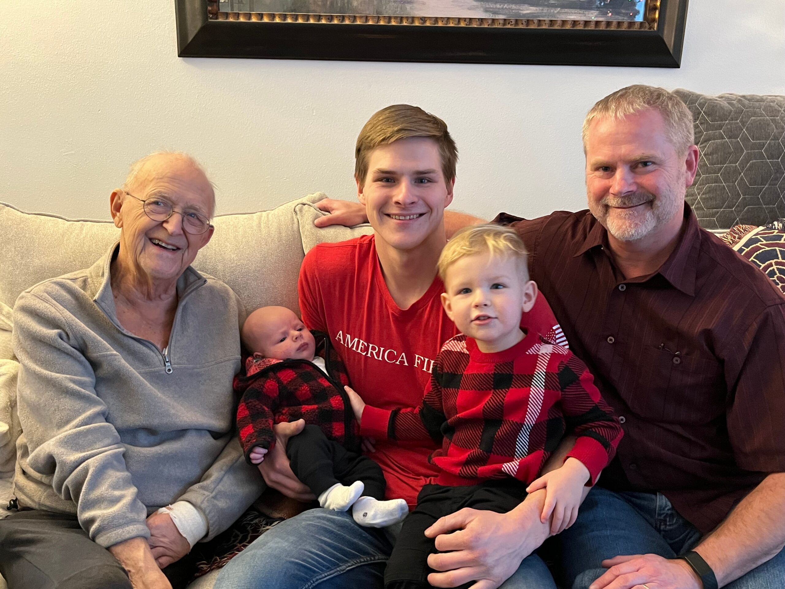 Family of five sitting together on a living-room sofa: grandfather on the left, teen in the middle, two young children, and a father on the right, all smiling.
