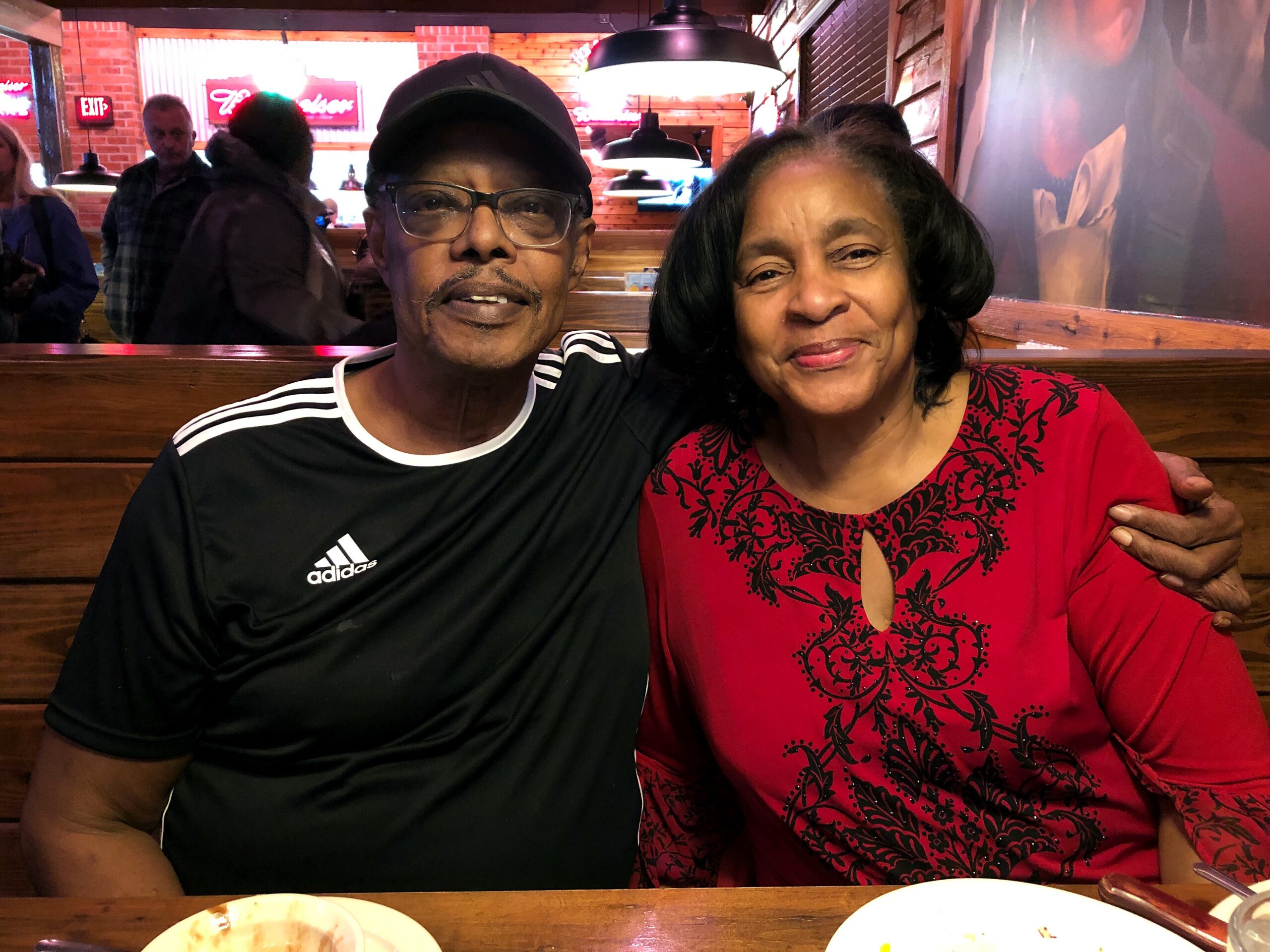 Smiling couple sitting together in a restaurant booth; man in a black Adidas shirt and cap, woman in a red patterned top, both looking at the camera.