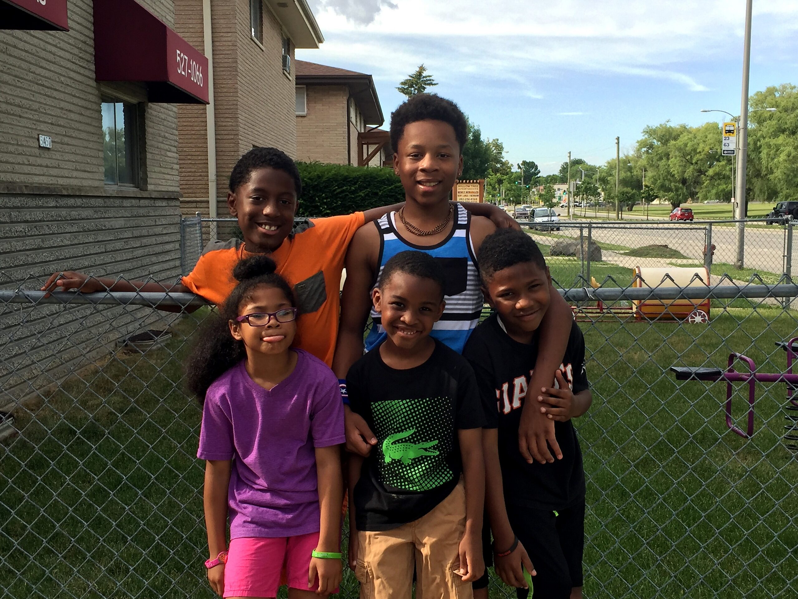 Six children posing and smiling behind a chain-link fence beside a brick building with a red awning.