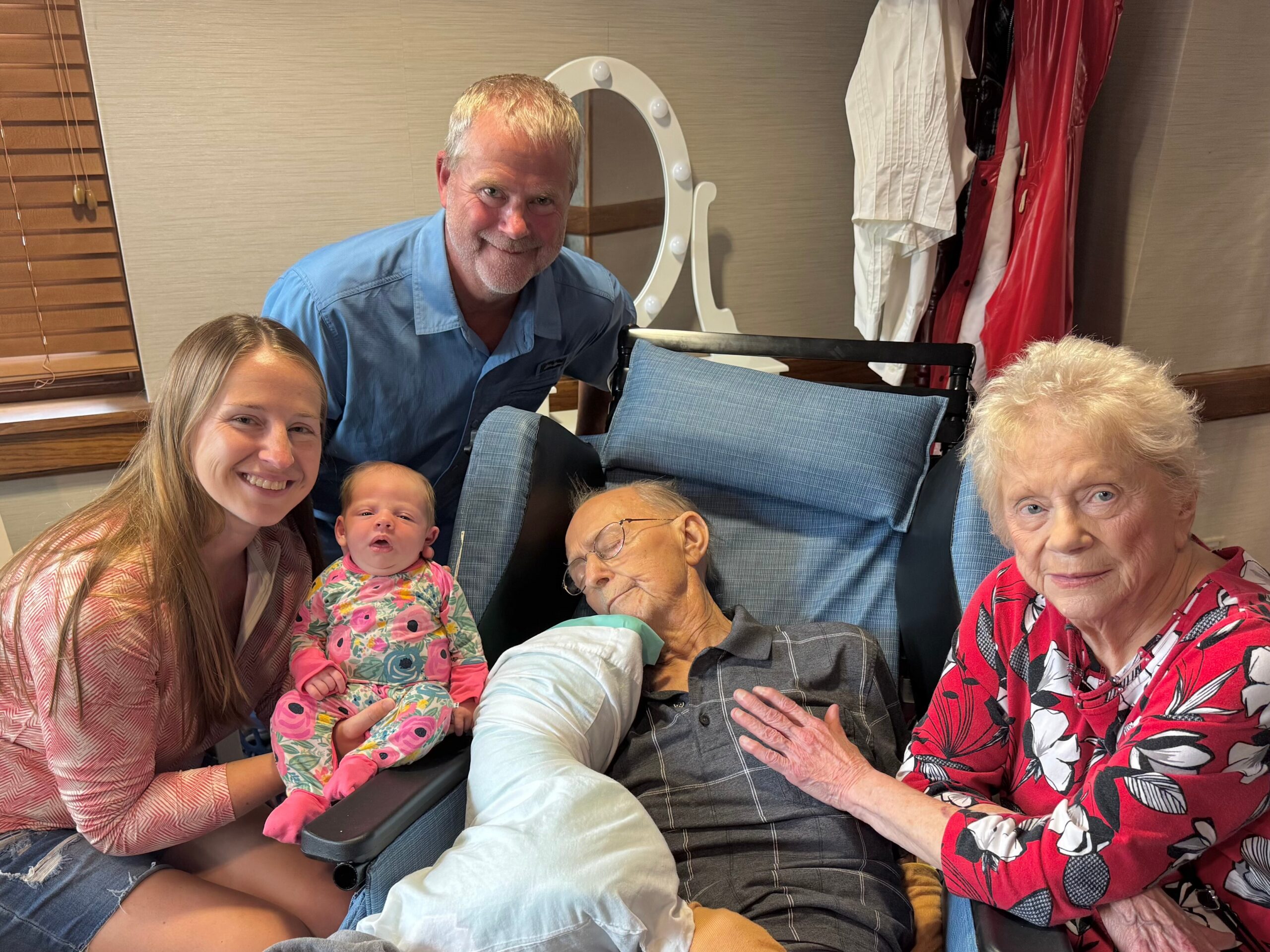 A group of people posing together in a room: a smiling young woman with a baby, an older woman in a wheelchair, and others nearby, with coats hanging in the background.