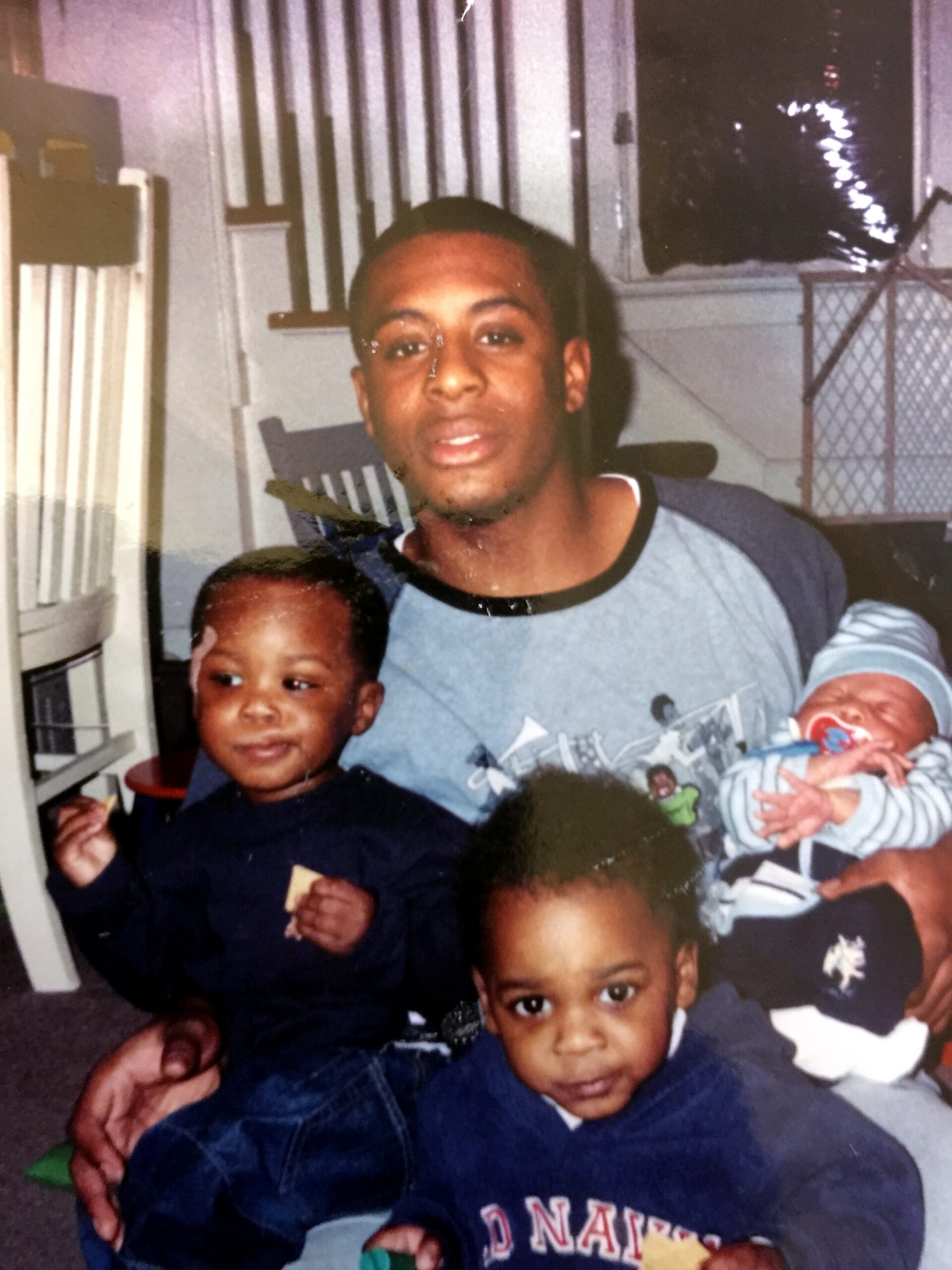 Man with two young children indoors, a toddler beside him and a baby in his arms, smiling for the photo.