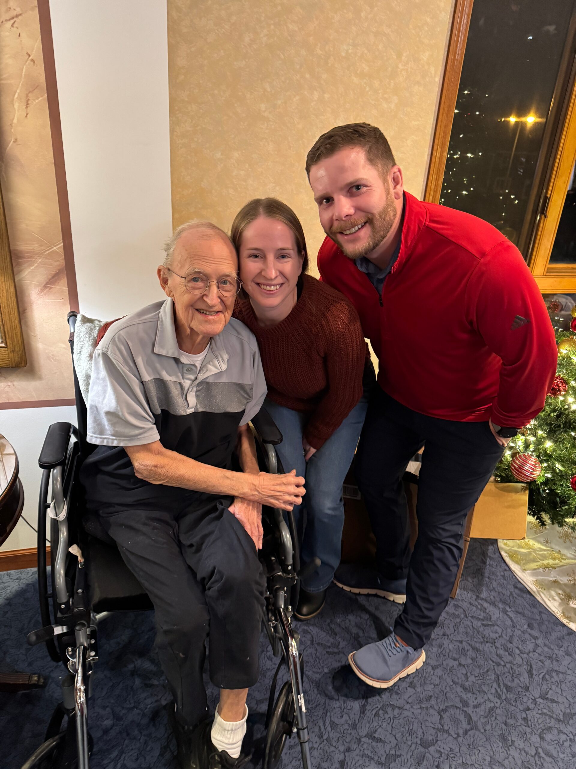 Three people posing for a photo: an elderly man in a wheelchair on the left, a smiling woman in the middle, and a man in a red jacket on the right, indoors near a Christmas tree.