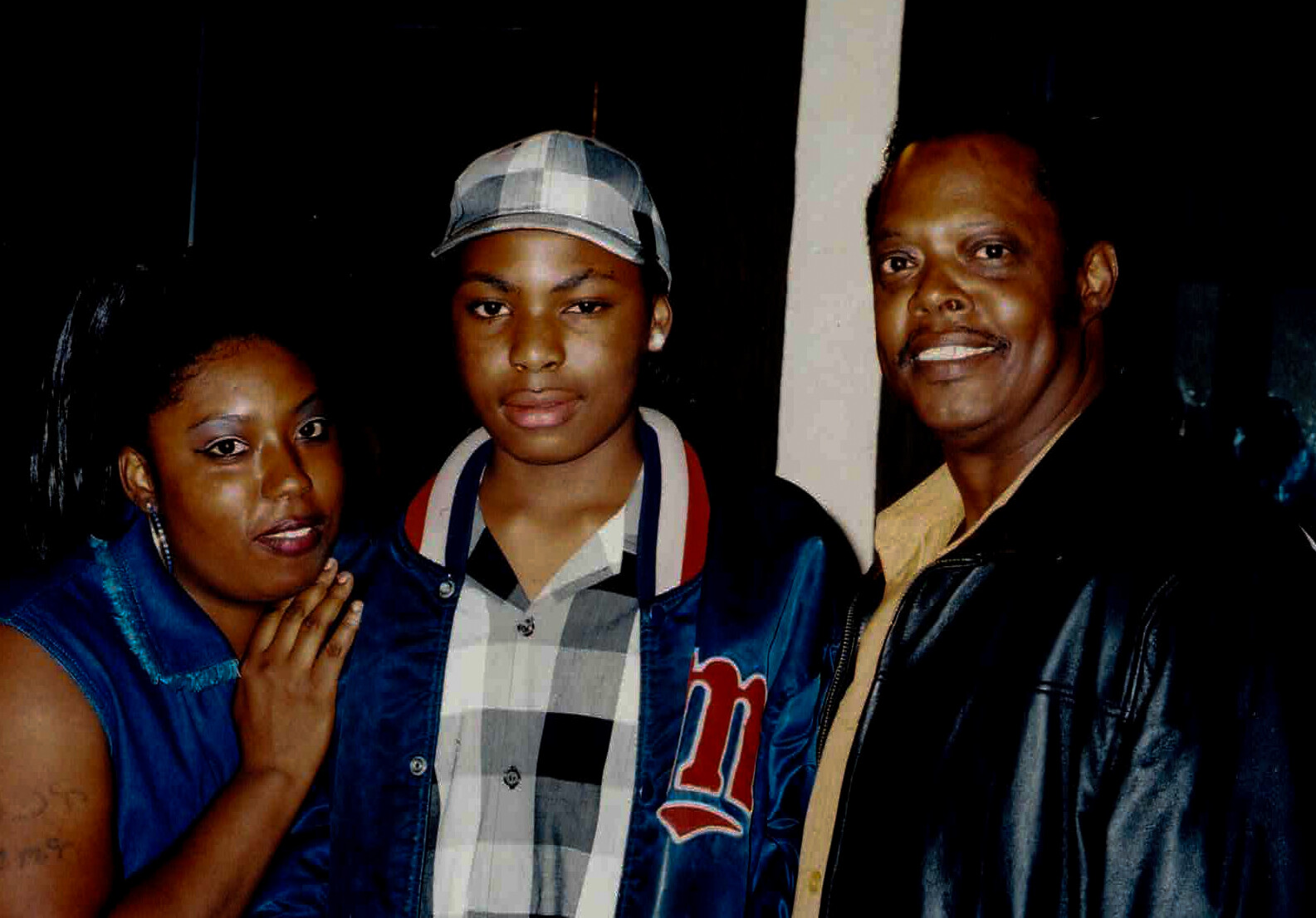 Three friends pose together indoors at night; left woman in a blue jacket, center person in plaid shirt and cap, right man in a leather jacket.