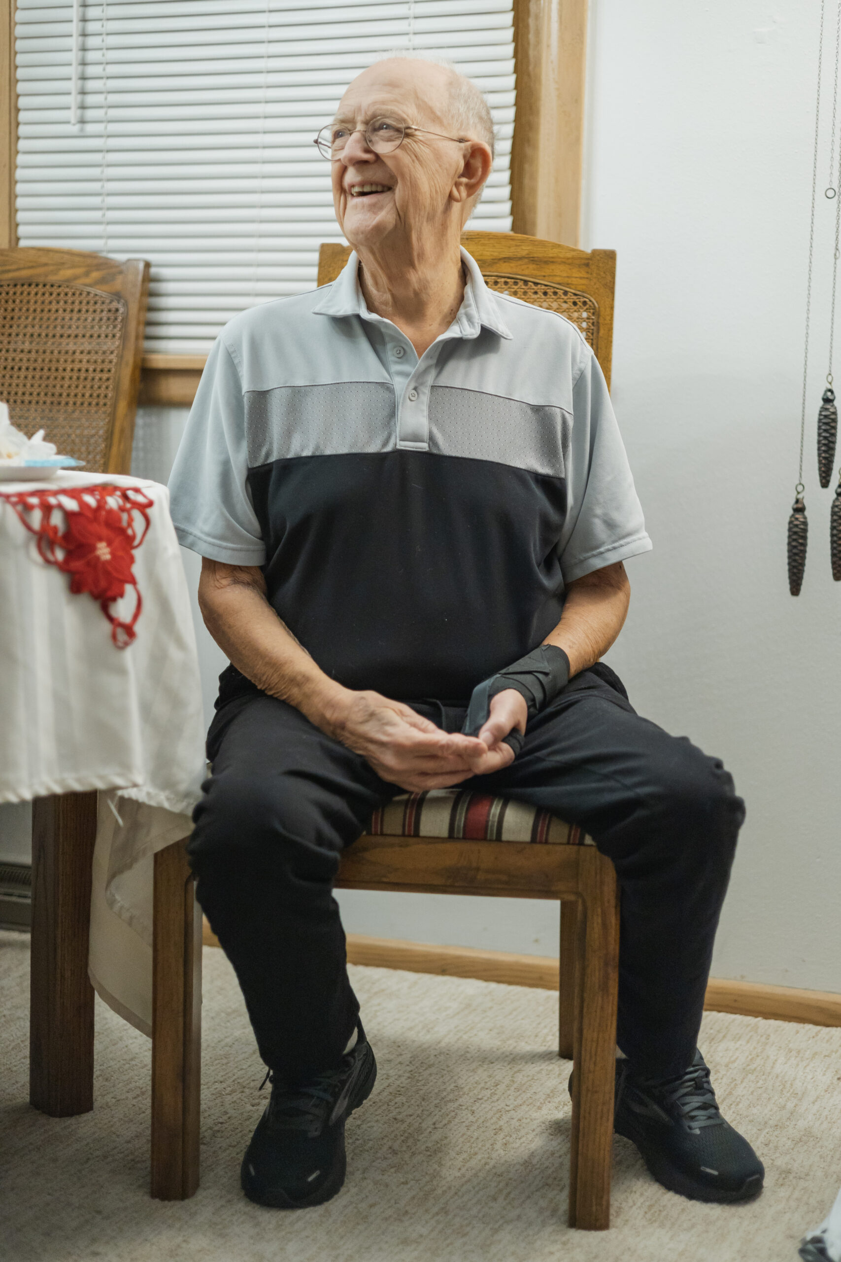 Older man with glasses smiling while seated on a wooden chair, wearing a light gray and navy polo and black pants in a home setting.