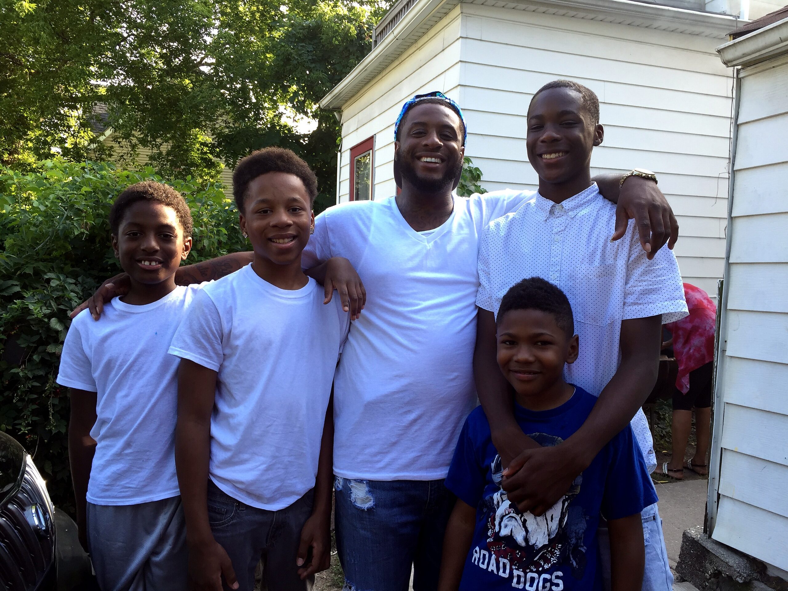 Group of smiling boys posing together outdoors in front of a white house and greenery behind them.