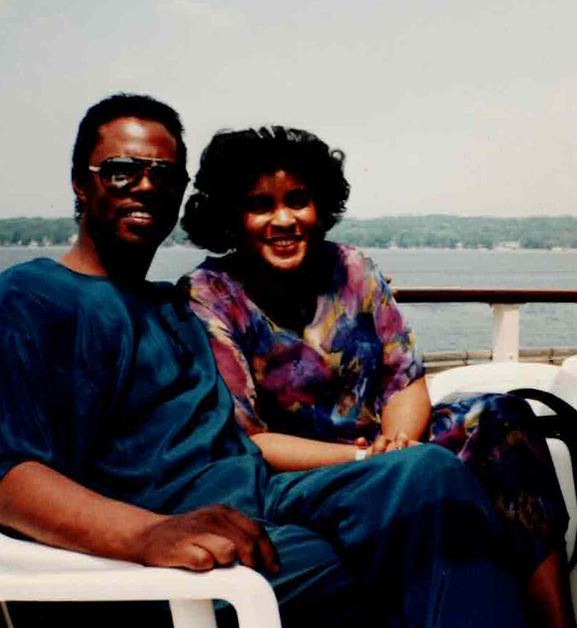 Couple sitting close together on a boat deck, smiling with water and shoreline in the background.