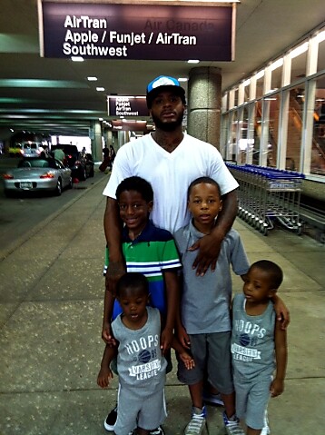 Man in a white T-shirt with four young boys posing at an airport terminal, luggage carts and signage in the background.