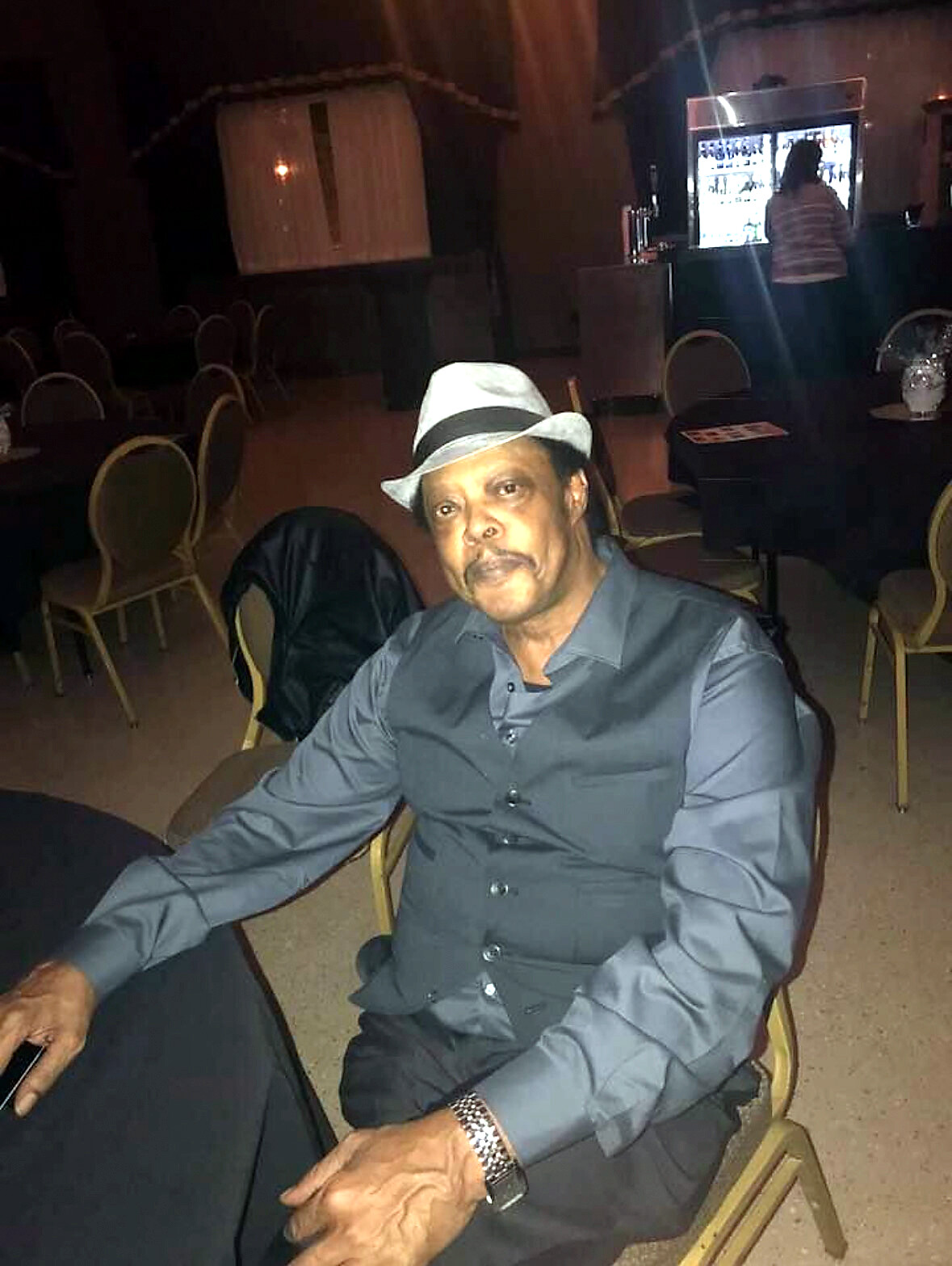 Man wearing a light gray fedora and gray shirt sits at a round table in a dim banquet hall during an event.