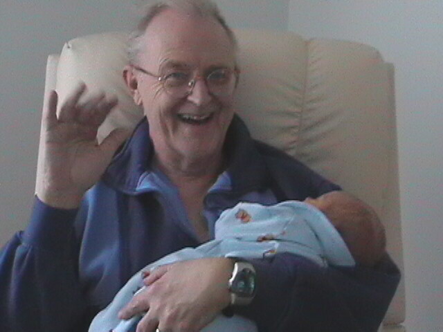 Older man with glasses sits in a chair, smiling and waving while cradling a newborn in a blue blanket.