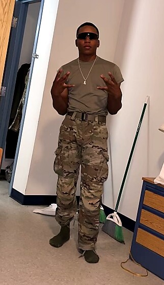 Young man in an olive T-shirt and camouflage cargo pants, wearing sunglasses and a chain, posing with hands raised in a hallway.