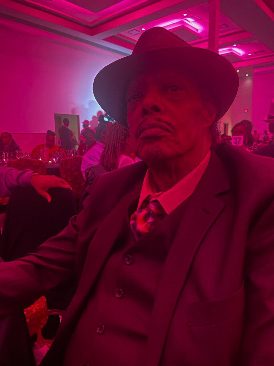 Older man in a fedora and dark suit at a pink-lit banquet hall, looking toward the camera.