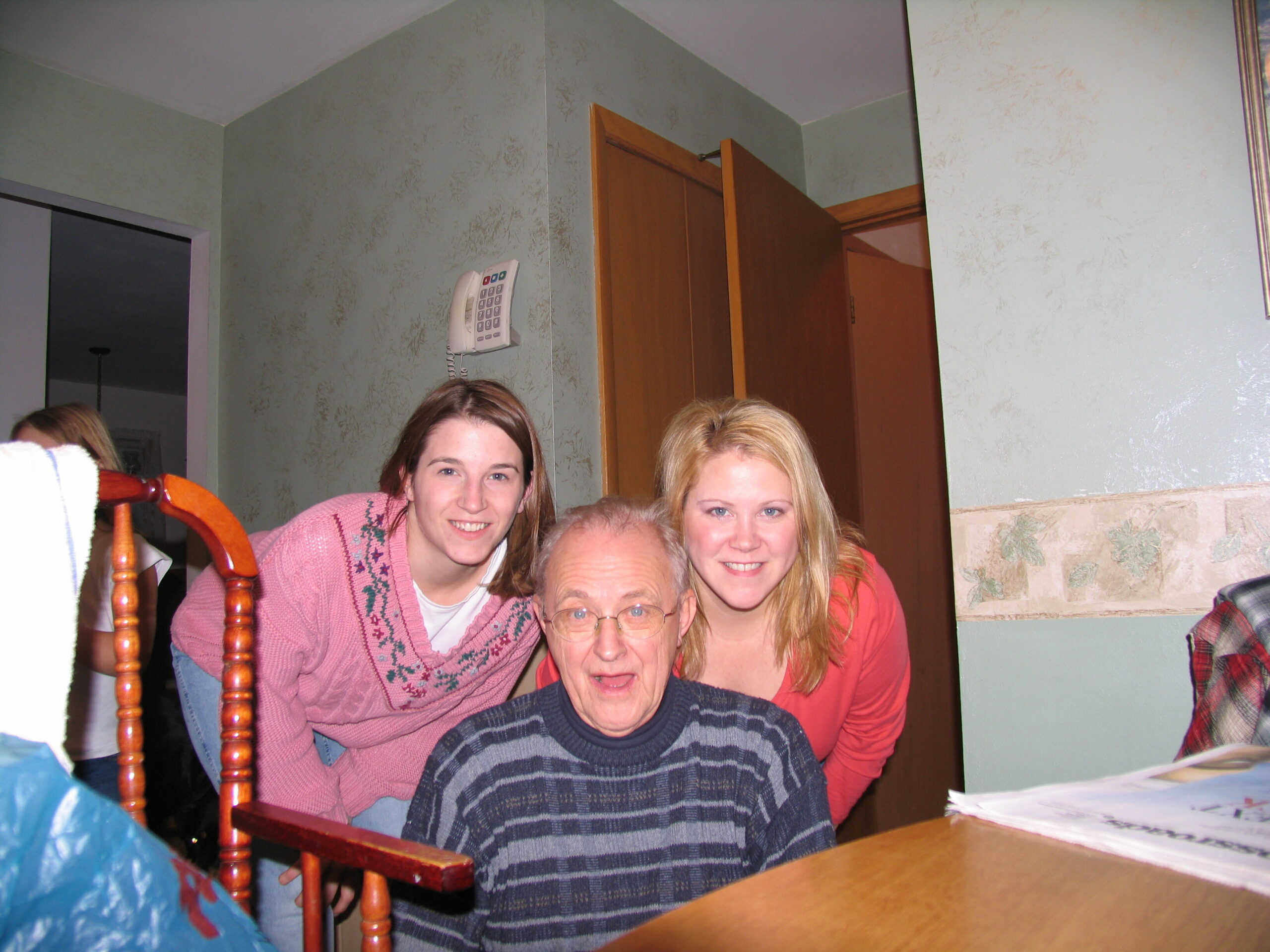 Group of four people posing for a photo indoors, with an older man in a striped sweater in the foreground and two smiling women behind him; a wall-mounted phone is on the pale green wall to the left.