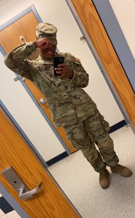 Person in camouflage military uniform taking a mirror selfie with a phone in a hallway.