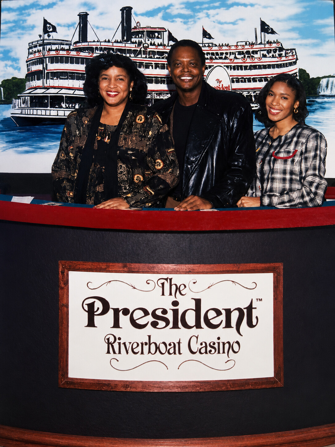 Three smiling people stand behind a red railing at The President Riverboat Casino, with a large riverboat backdrop behind them and its logo visible behind the group.