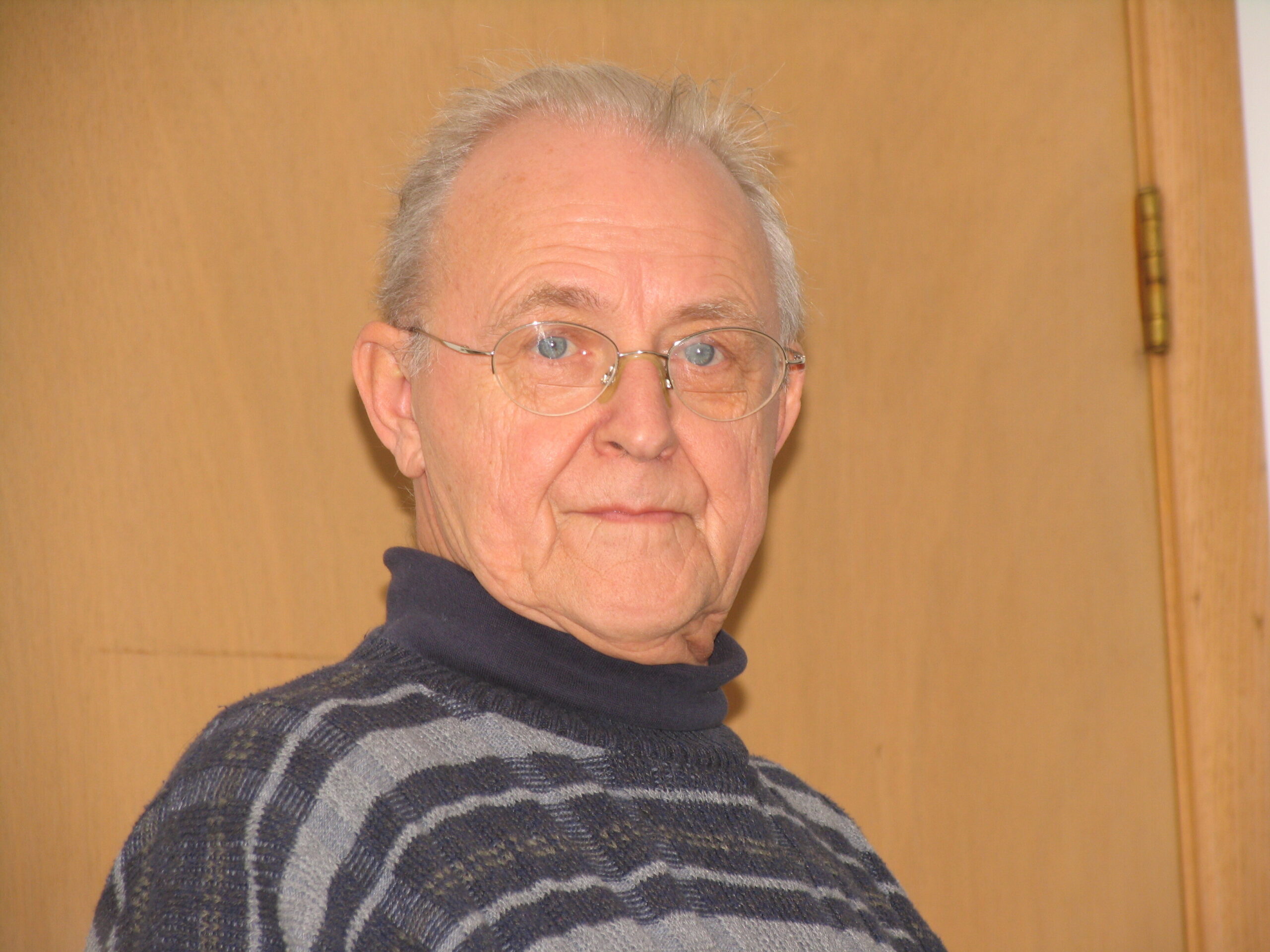 Older man with glasses wearing a dark, striped sweater, positioned indoors against a wooden door, looking slightly to the right.