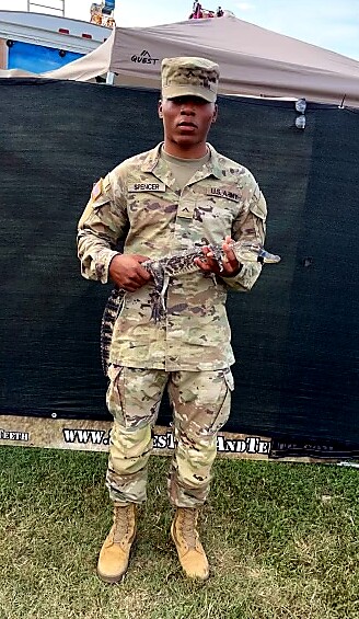 U.S. Army soldier in camouflage holding a snake at an outdoor event, with tents in the background.