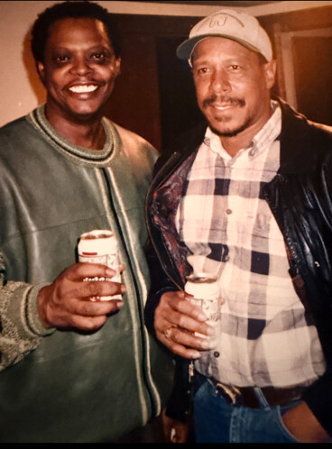 Two men posing indoors, smiling and holding drinks; left man in a green jacket, right man in a plaid shirt and cap.